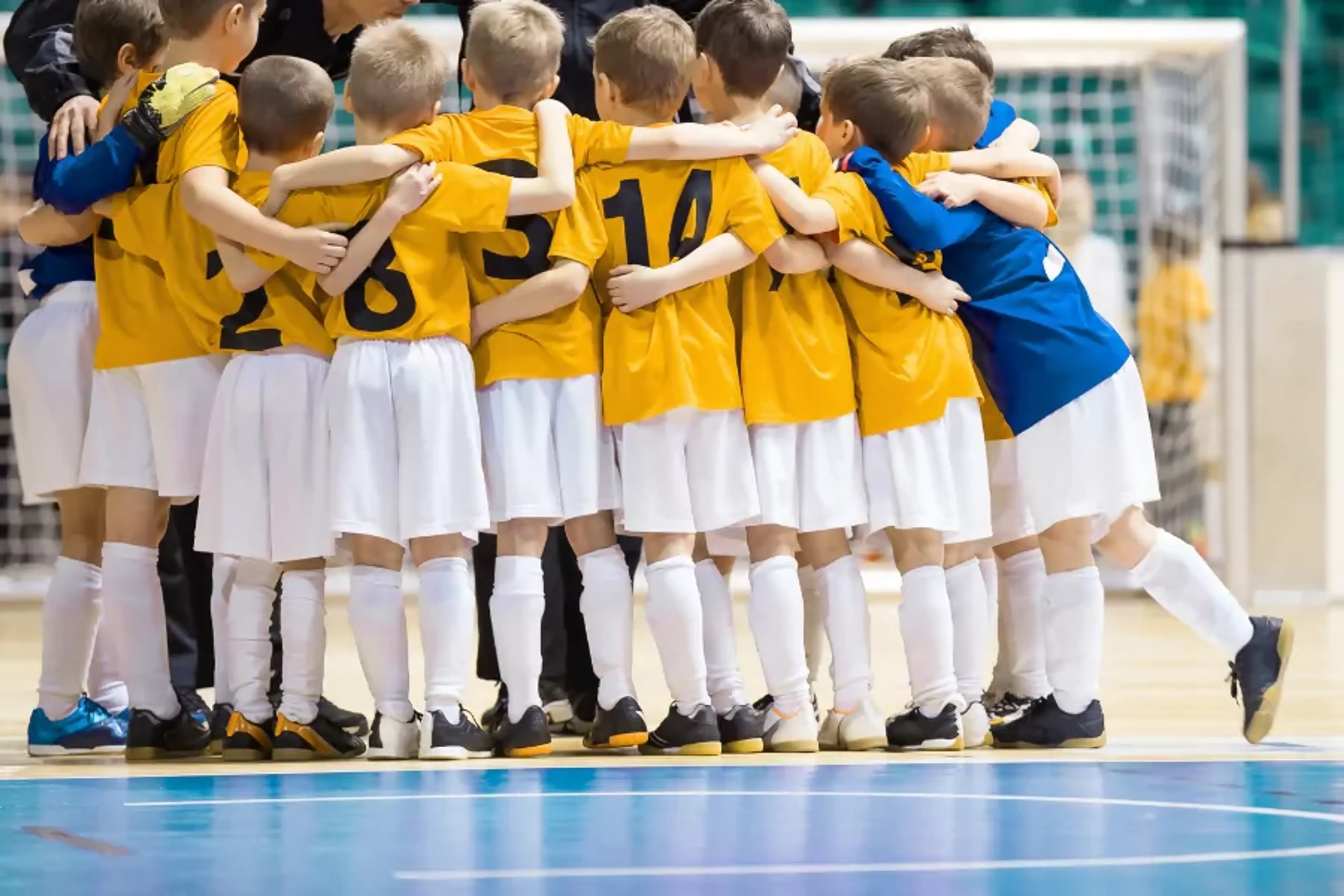 En grupp barn i gula matchtröjor och vita shorts står tätt tillsammans i en cirkel på en futsalarena. Ett barn i blå tröja är en del av gruppen vid sidan.