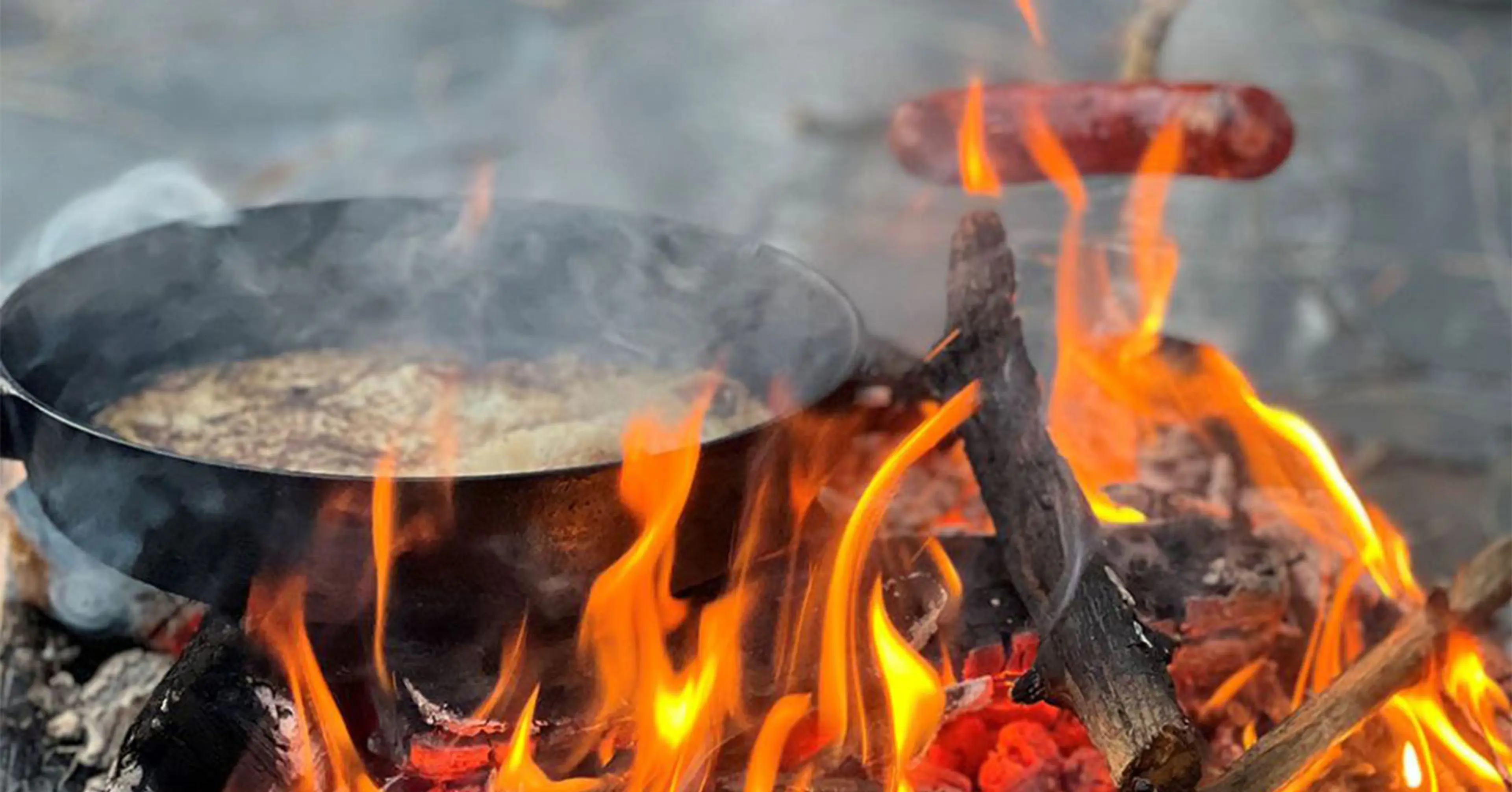 En stekpanna över öppen eld där något steks, och en korv som grillas på en pinne ovanför lågorna. Träbitar brinner och skapar en sprakande lägereld.