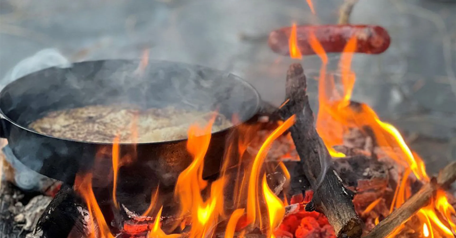 En stekpanna över öppen eld där något steks, och en korv som grillas på en pinne ovanför lågorna. Träbitar brinner och skapar en sprakande lägereld.