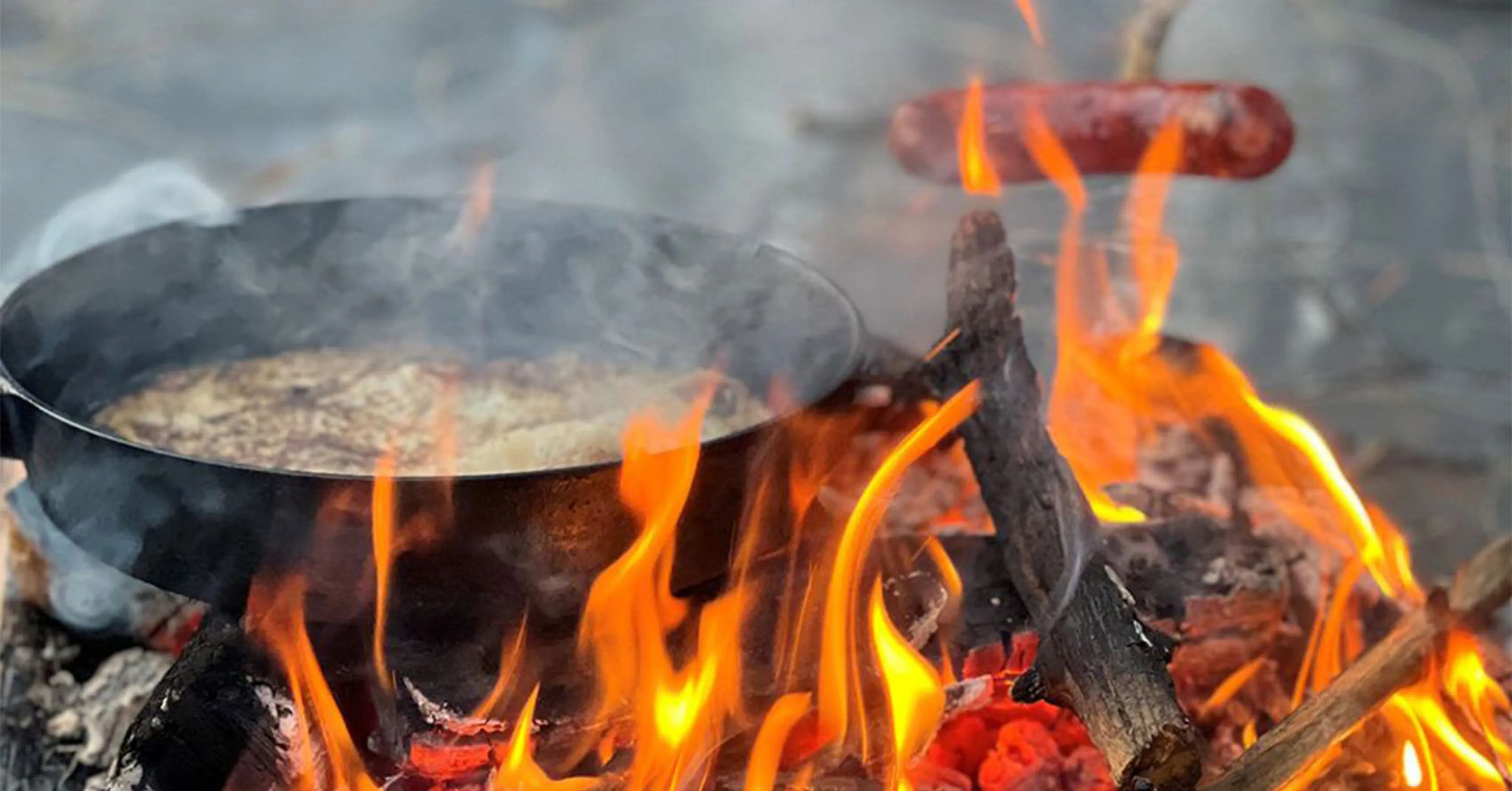 En stekpanna över öppen eld där något steks, och en korv som grillas på en pinne ovanför lågorna. Träbitar brinner och skapar en sprakande lägereld.