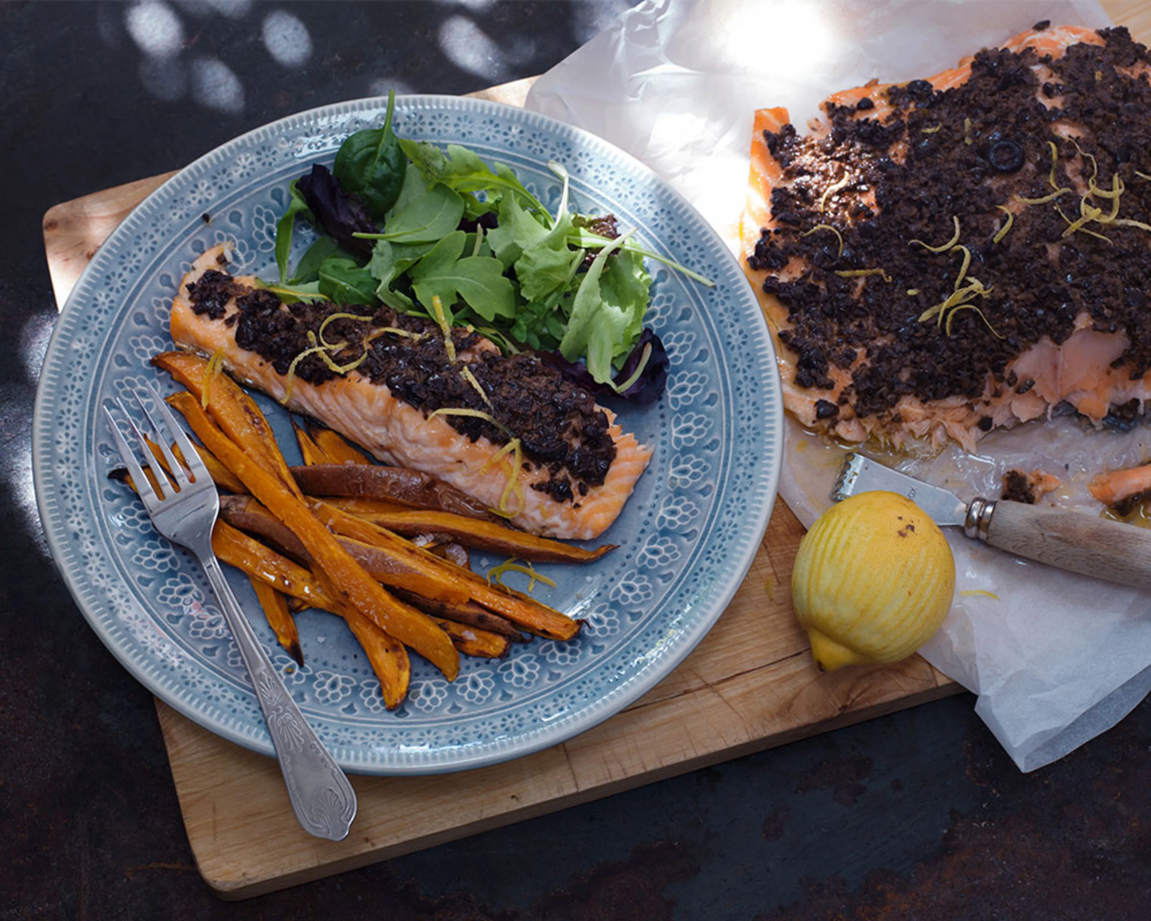 En portion ugnsbakad lax med tapenade, sötpotatisstavarna och en fräsch grönsallad på en blåmönstrad tallrik, bredvid ligger en bit lax med tapenade och en citron.