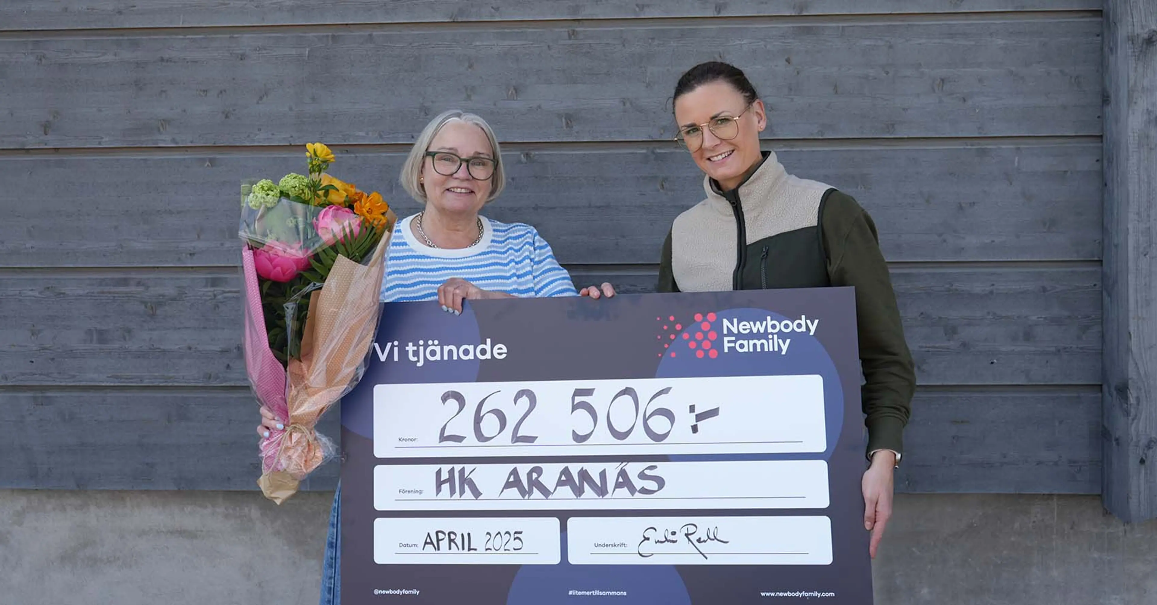 Två kvinnor håller upp en stor check med texten "262 506 kr" och "HK Aranas" skrivna på, ena kvinnan har en blombukett i handen. Bakgrunden är en grå trävägg.
