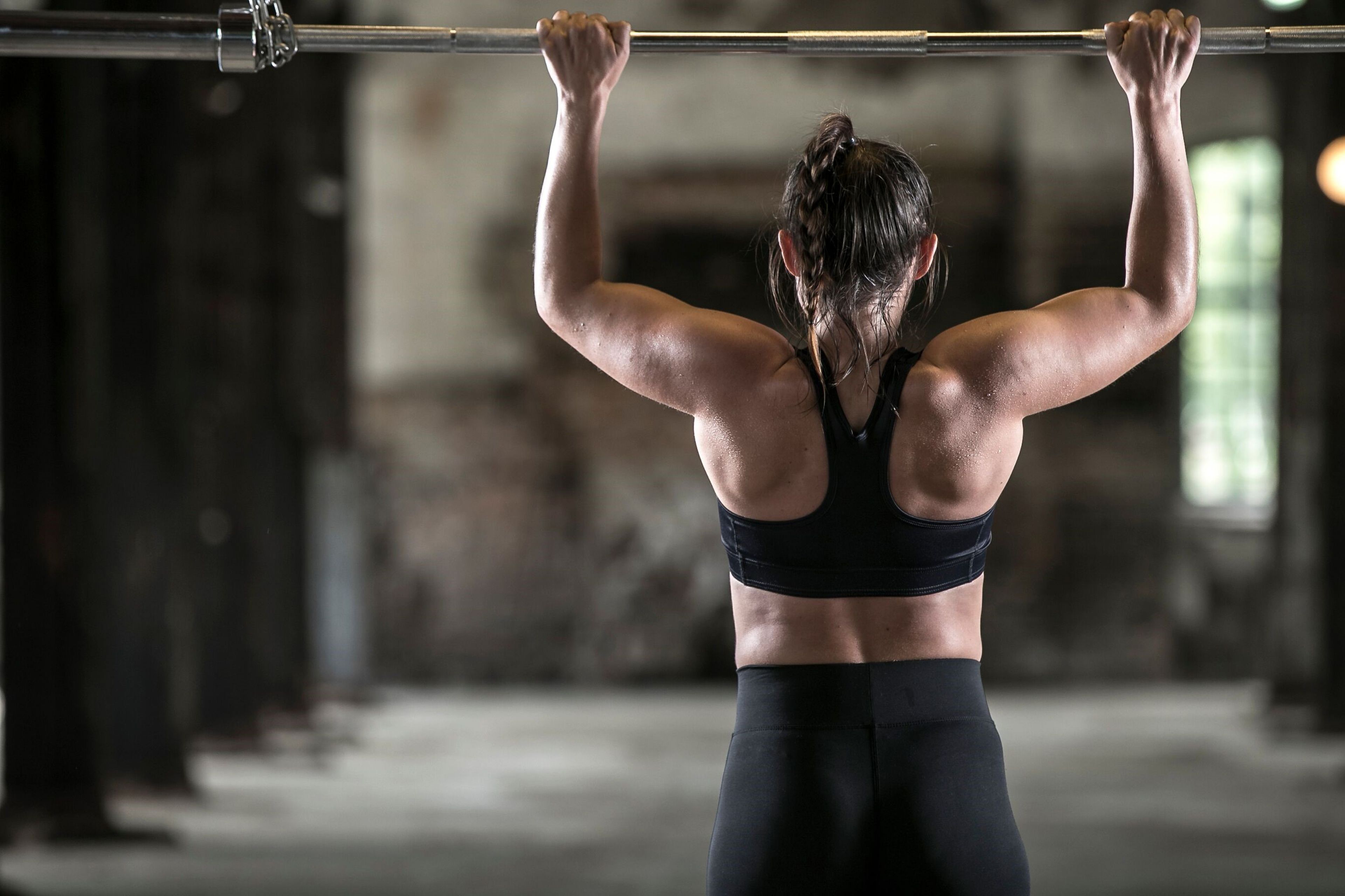 Kvinna i sport-bh och svarta träningsleggings som gör ett pull-up grepp på en skivstång. Bakgrunden är en industrilokal med mjukt ljus.