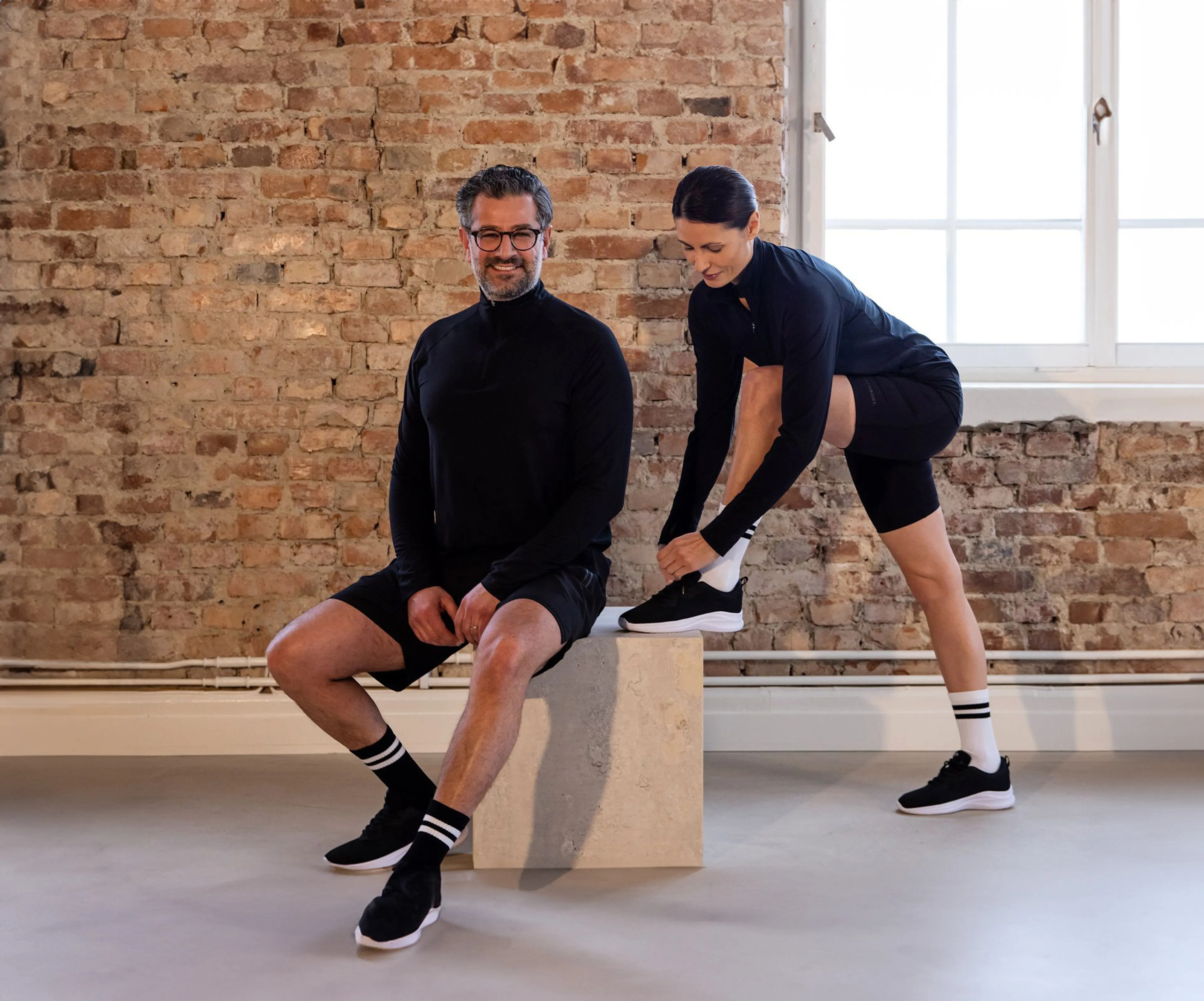 En man och en kvinna bär svarta långärmade tröjor med dragkedja, den ena står och ler medan den andra knyter sina skor. Bakgrunden är en vägg av rå tegelsten och ett stort fönster släpper in ljus.