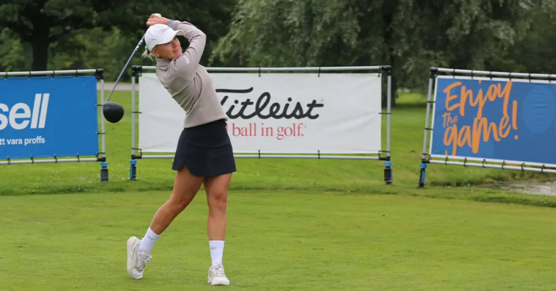 En person klädd i ljus tröja och mörk kjol svingar en golfklubba på en grön golfbana. Bakom syns reklamskyltar med texten "Enjoy the game!" och "Titleist".