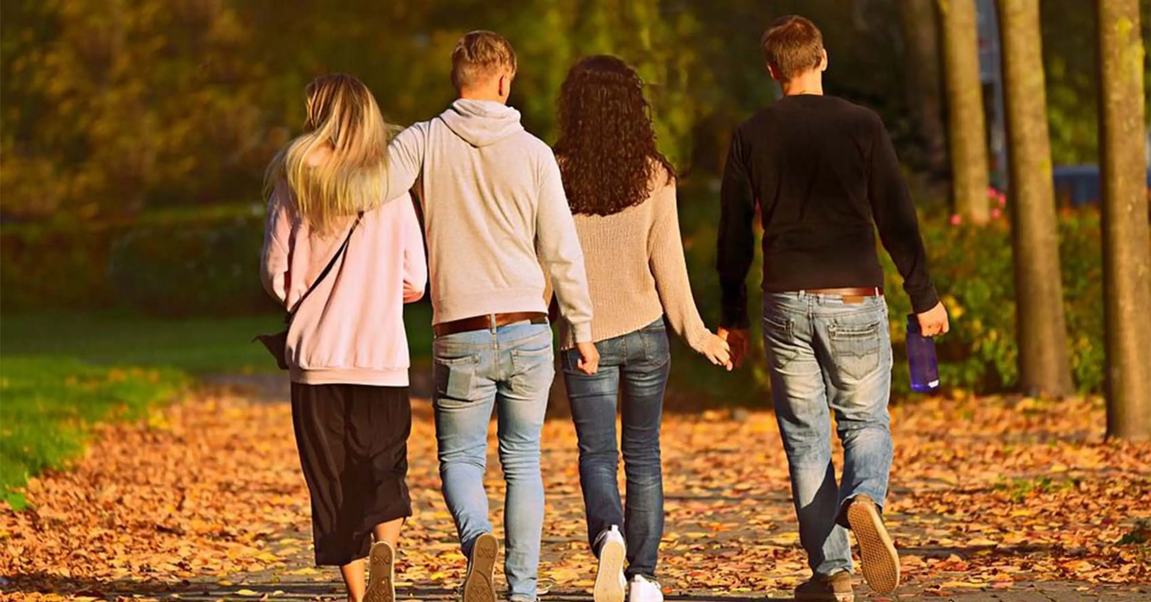 Fyra personer går tillsammans på en lövbeklädd stenlagd stig bland träd på hösten. Två håller varandra i handen medan de promenerar bort från kameran.