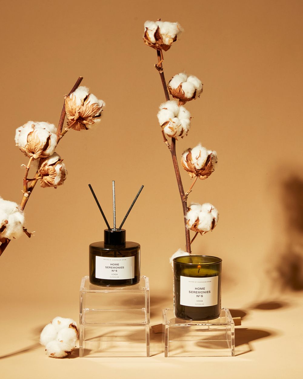 En doftspridare och ett doftljus i mörkt glas med vita etiketter, omgivna av bomullsgrenar mot en varm beige bakgrund.
