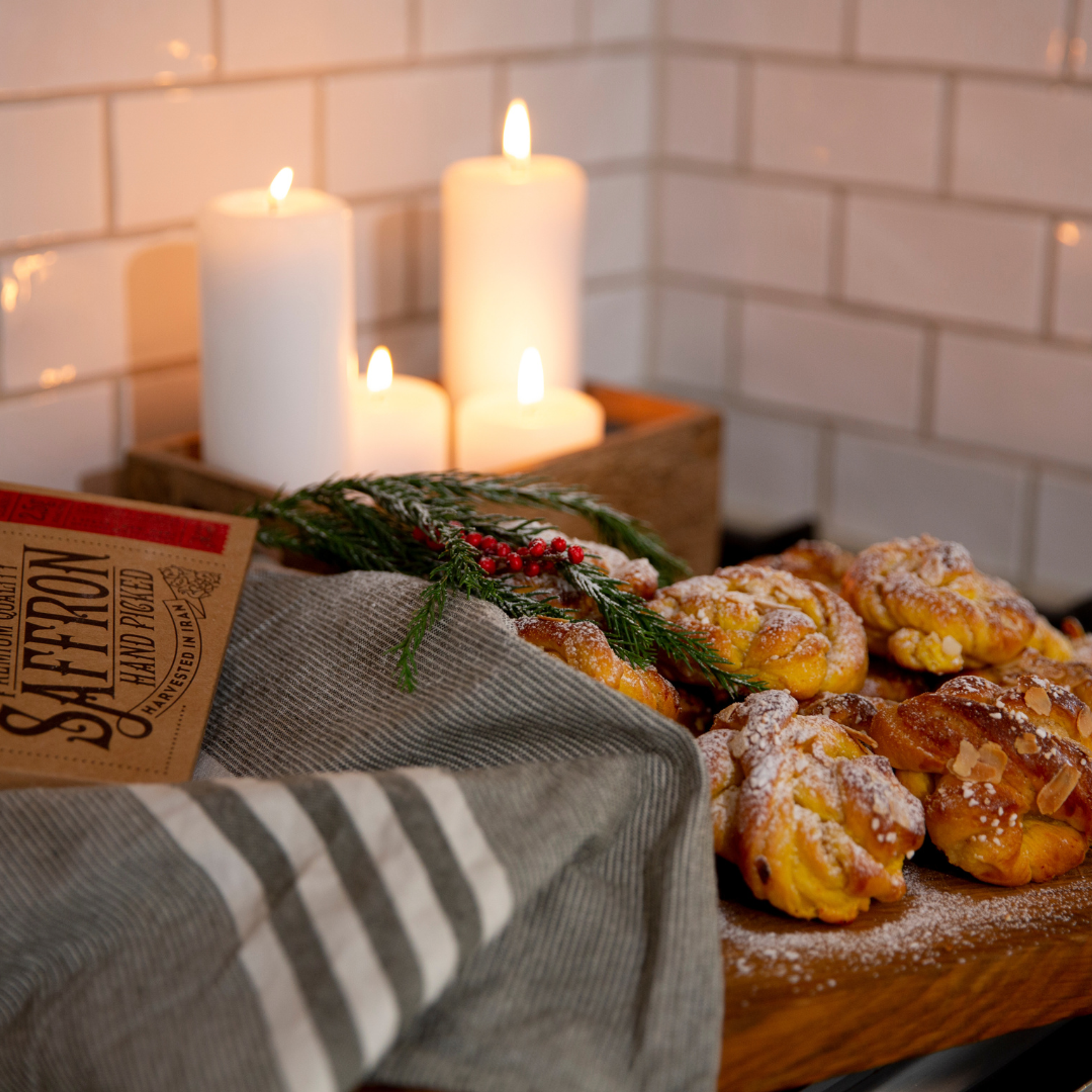 Färska saffransbullar med florsocker och mandelspån på ett träfat, dekorerade med granris och röda bär. Fyrkantiga vita blockljus lyser i bakgrunden mot vitt kakel.