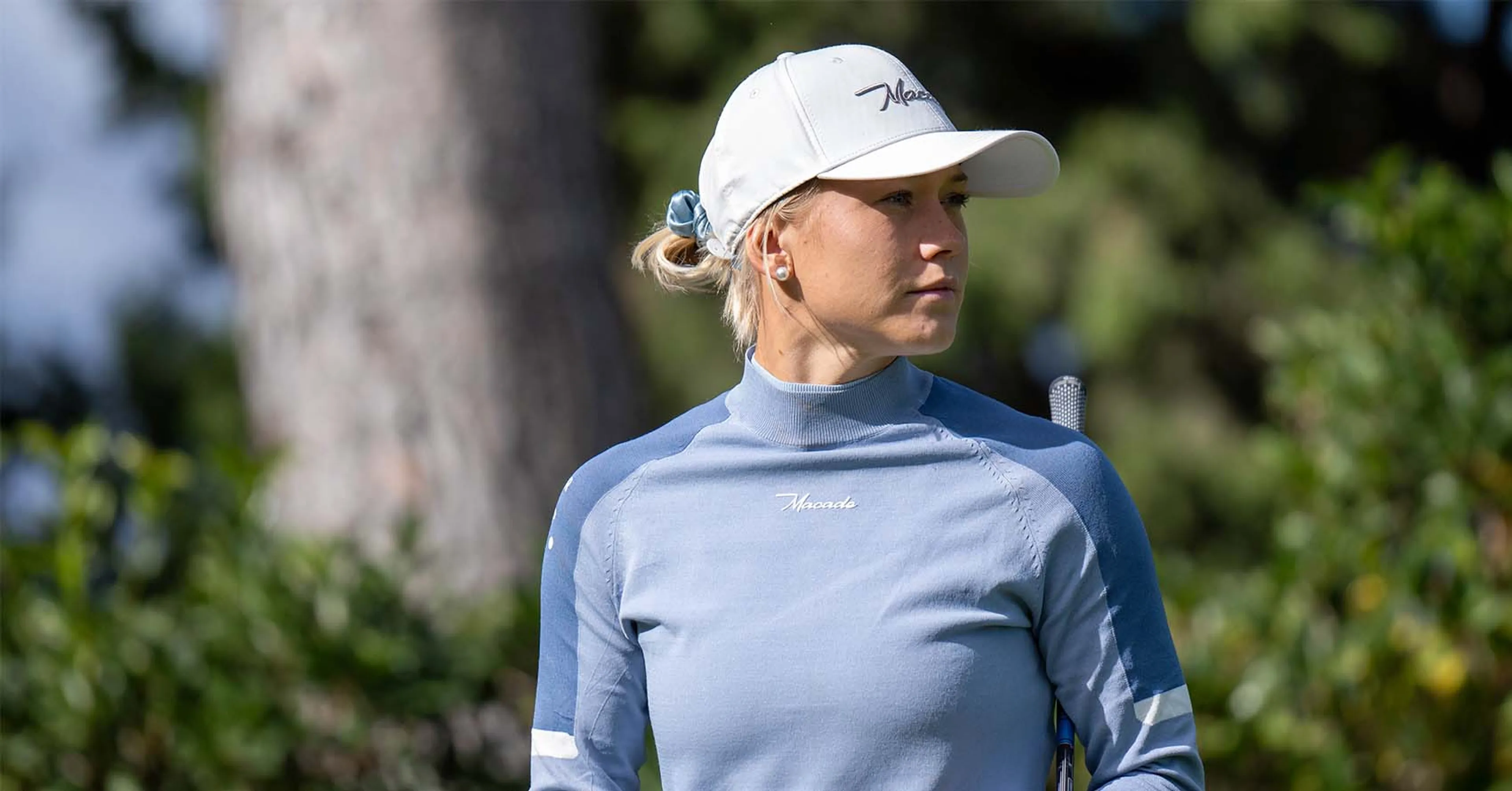 En kvinna i ljusblå golftröja och vit keps står i en grönskande utomhusmiljö med en golfklubba vid sidan. Tröjan har en hög hals och en enkel, sportig design.