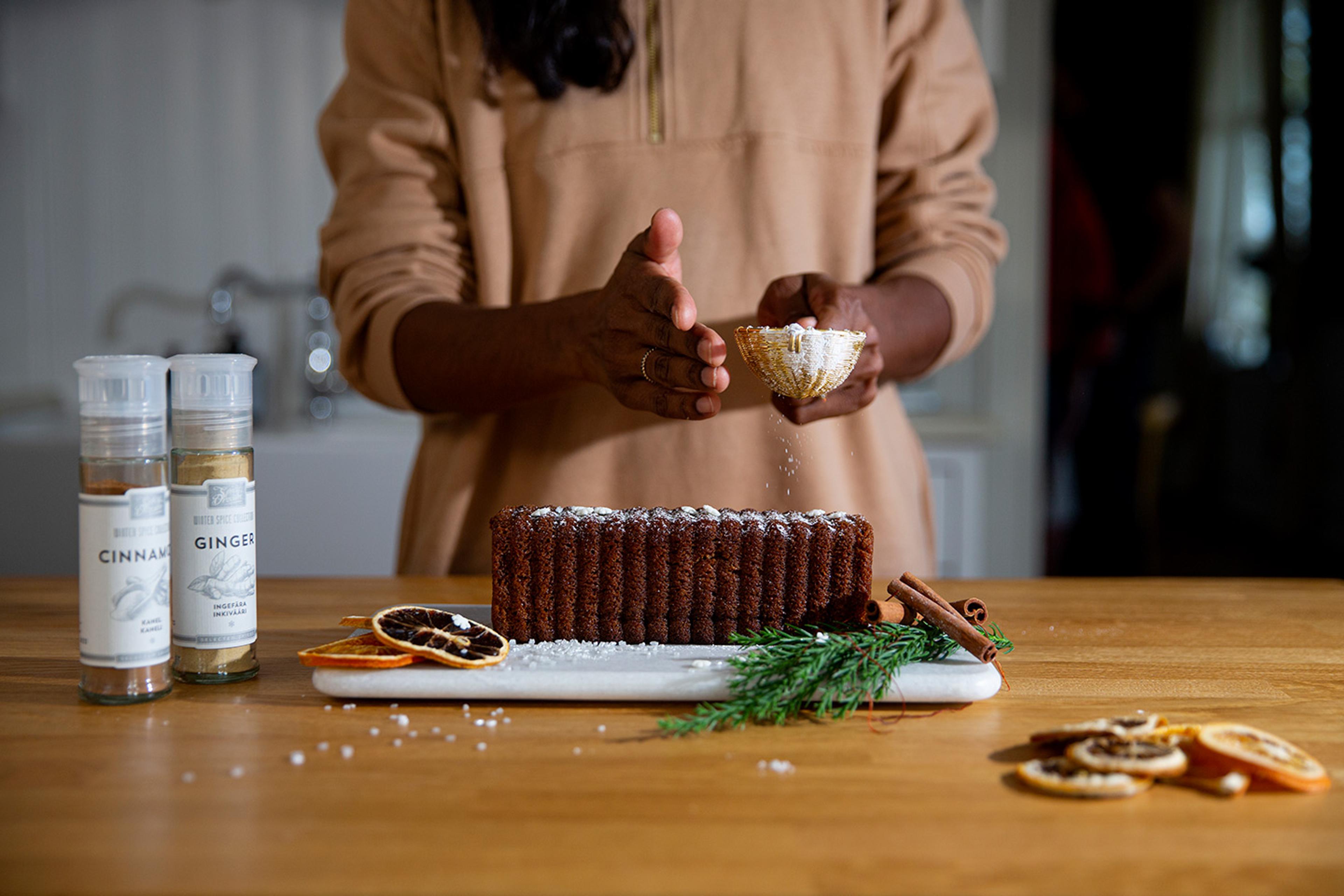En hand strör florsocker över en avlång kryddkaka på ett träbord, omgiven av torkade apelsinskivor, kanelstänger och färsk granris. Två kryddburkar med kanel och ingefära står bredvid.