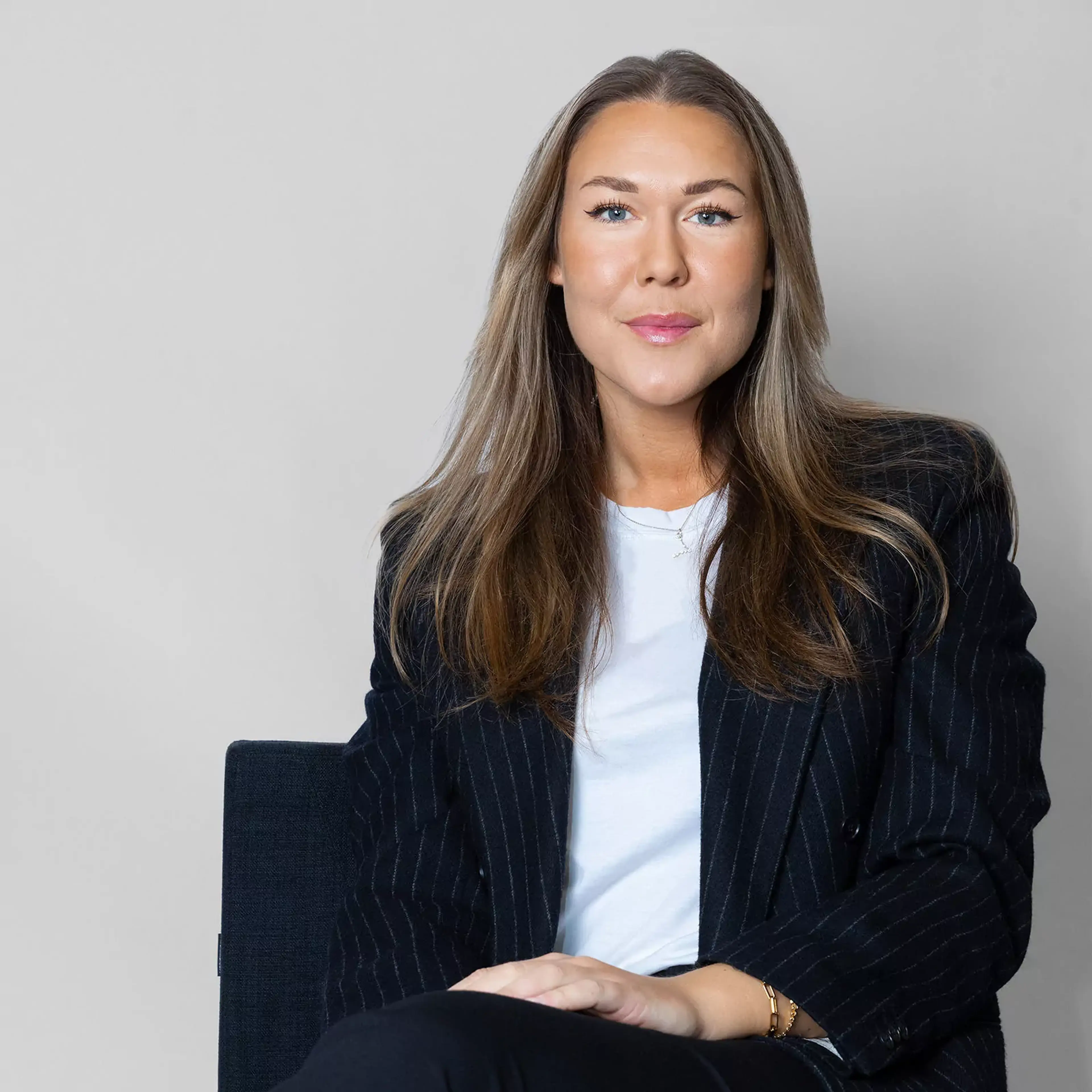 En kvinna med långt ljust hår bär en vit t-shirt och en mörkblå, randig kavaj medan hon sitter på en stol mot en ljusgrå bakgrund.