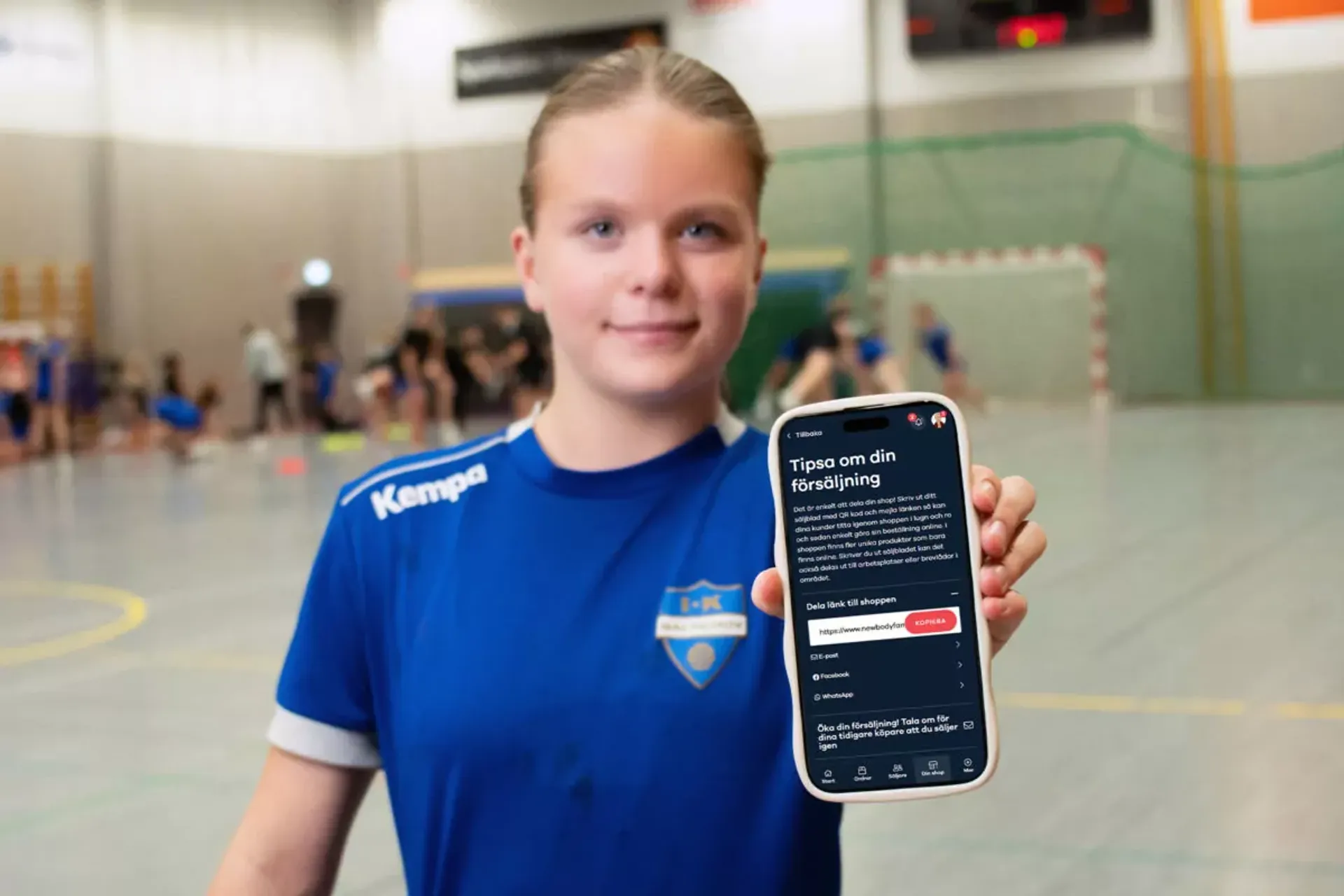 En ungdom i blå handbollströja håller upp en mobiltelefon med försäljningsappen öppen. Bakgrunden visar en handbollsplan där andra spelare är i rörelse.