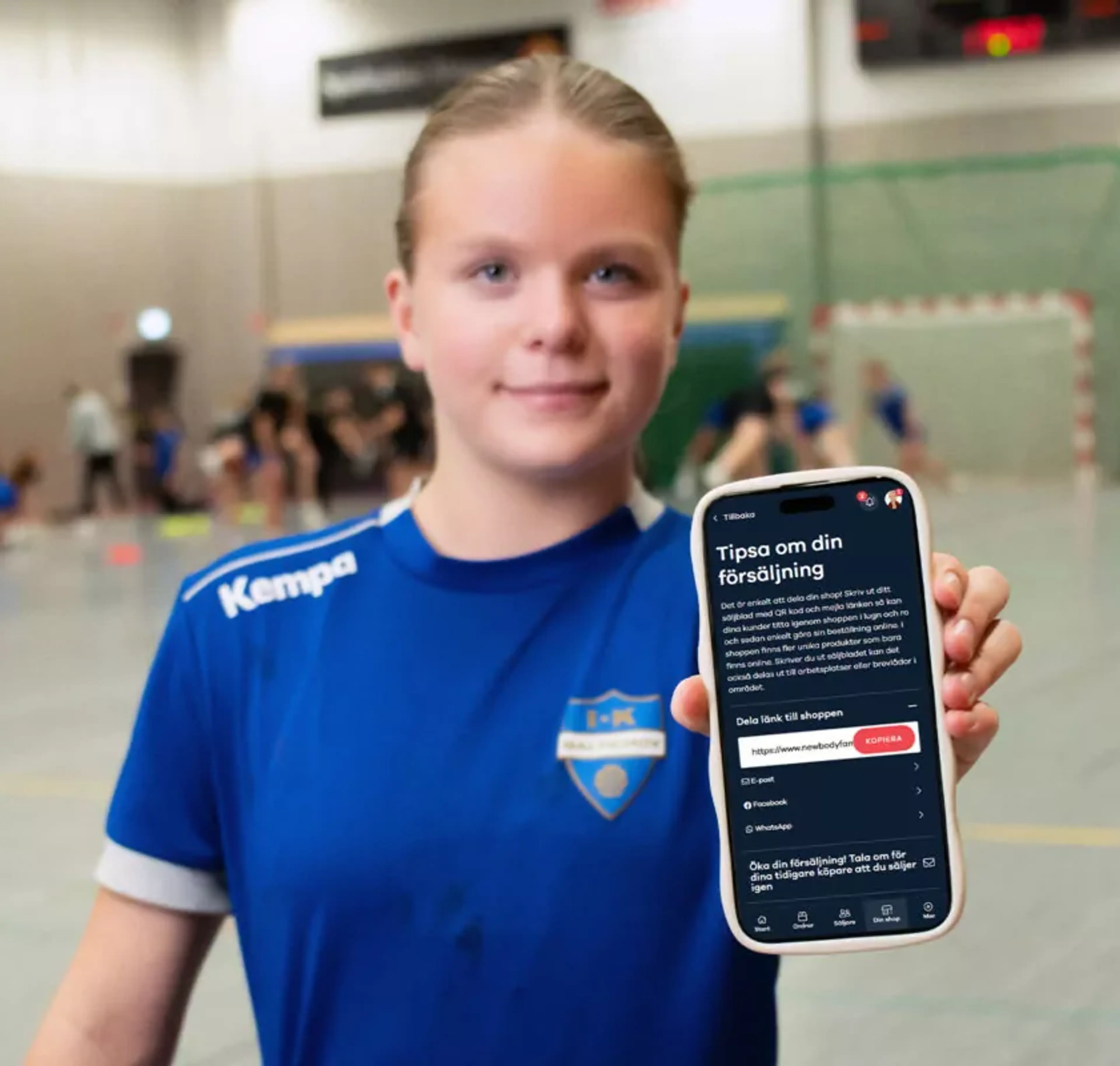 En ungdom i blå handbollströja håller upp en mobiltelefon med försäljningsappen öppen. Bakgrunden visar en handbollsplan där andra spelare är i rörelse.
