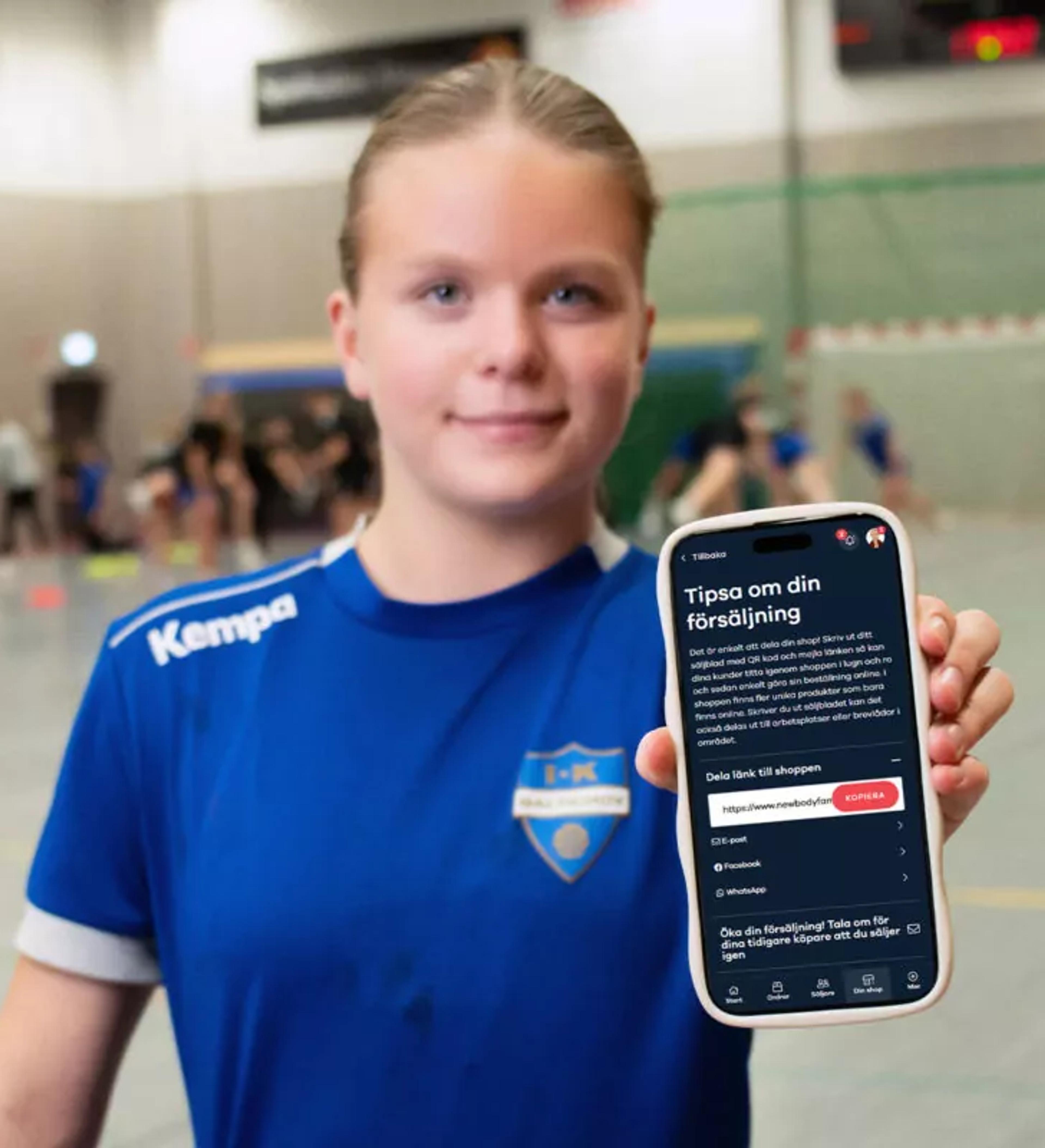 En ungdom i blå handbollströja håller upp en mobiltelefon med försäljningsappen öppen. Bakgrunden visar en handbollsplan där andra spelare är i rörelse.