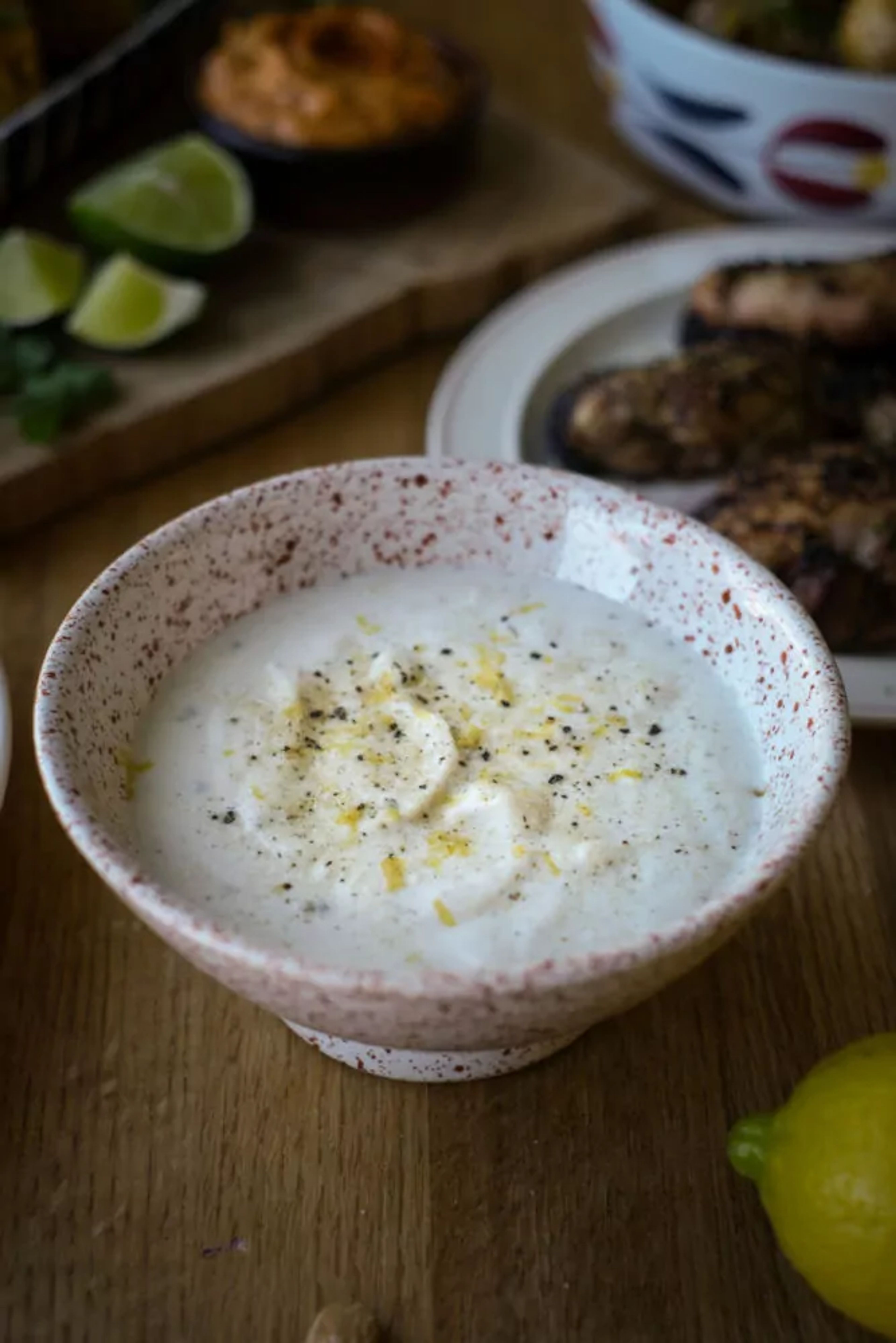 En skål med en krämig vit sås toppad med svartpeppar och citronskal. I bakgrunden syns limeklyftor och grillade bitar på en tallrik.
