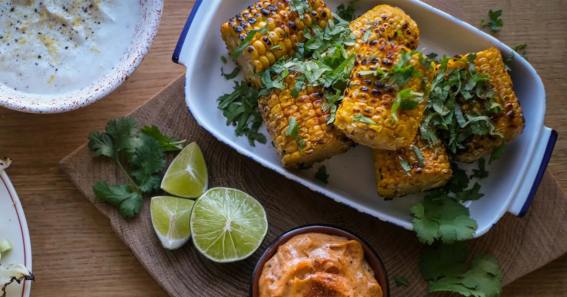 Grillade majskolvar med chilipulver och koriander i en ugnsform, serverade med en krämig, kryddig dipp och limeklyftor på ett träfat.