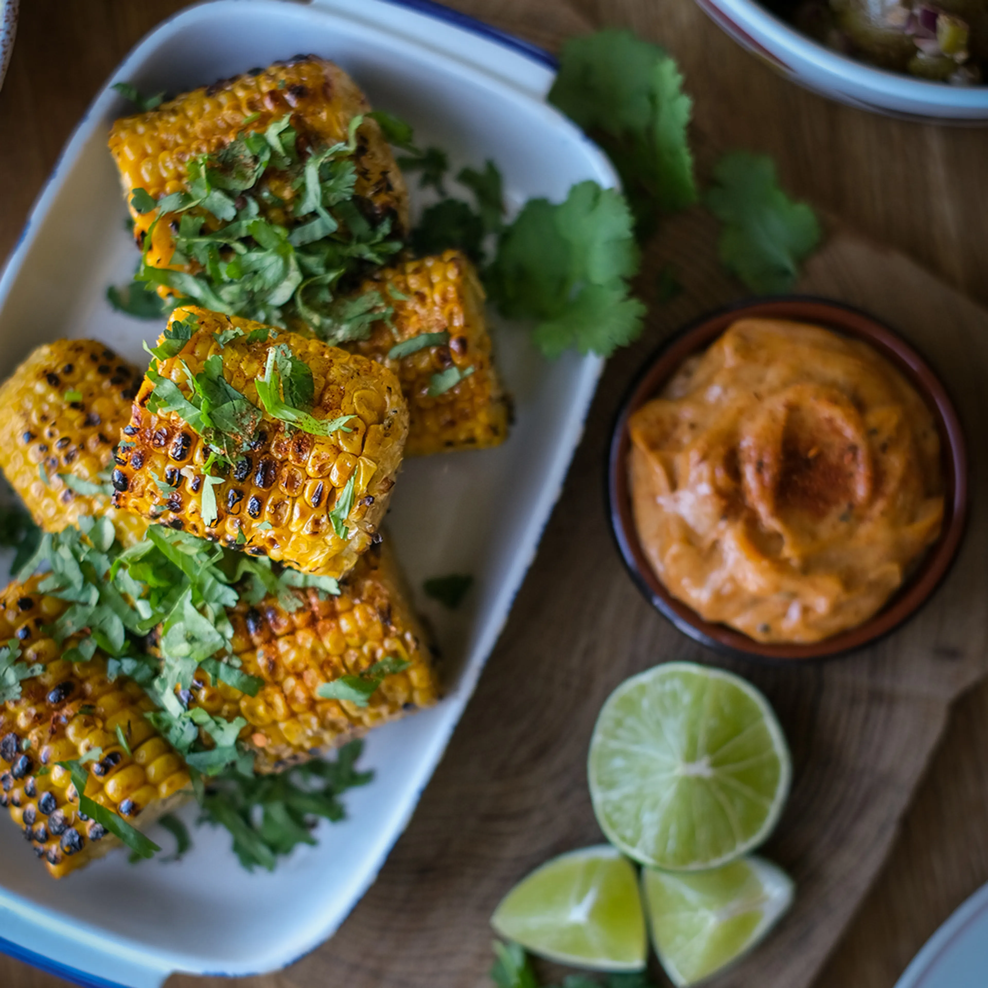 Grillade majskolvar med kryddig rubb och färsk koriander på en vit tallrik, serverade med limeklyftor och en krämig dippsås i en skål.