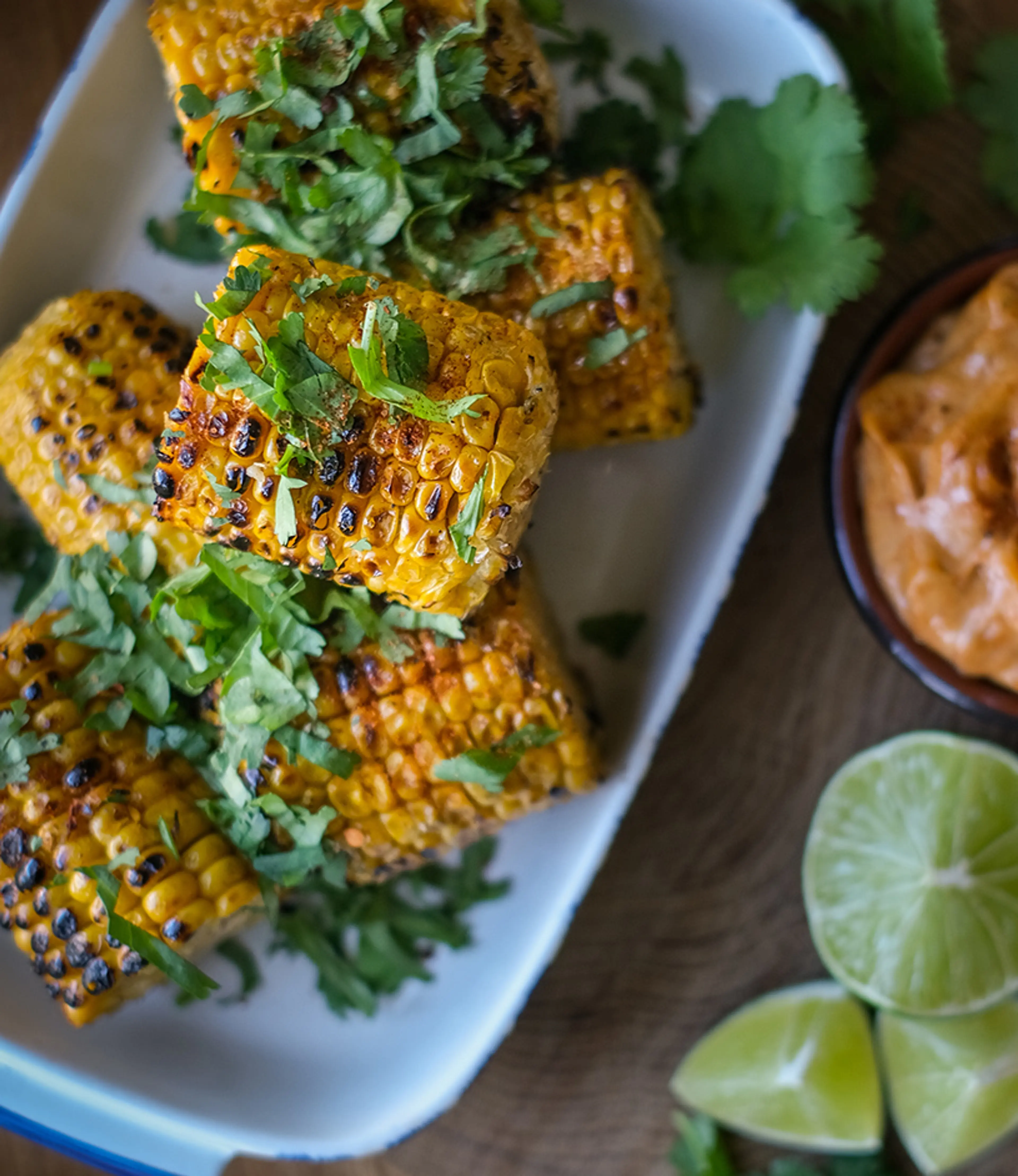 Grillade majskolvar med kryddig rubb och färsk koriander på en vit tallrik, serverade med limeklyftor och en krämig dippsås i en skål.