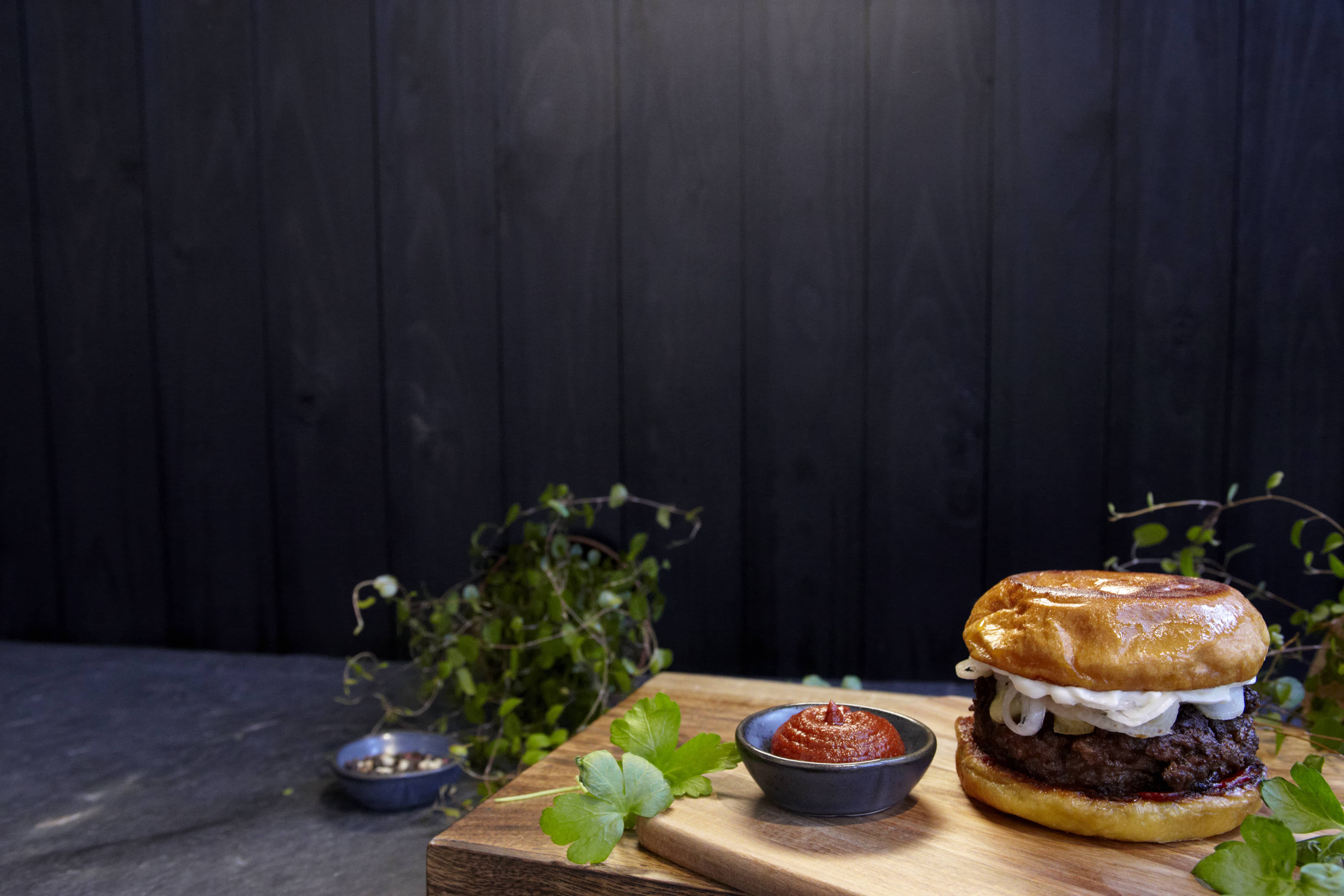 En saftig hamburgare med stekt lök i ett gyllene, mjukt hamburgerbröd står på en skärbräda tillsammans med en skål kryddig rödpaprikasås och färska persiljeblad. Mörk träbakgrund och gröna