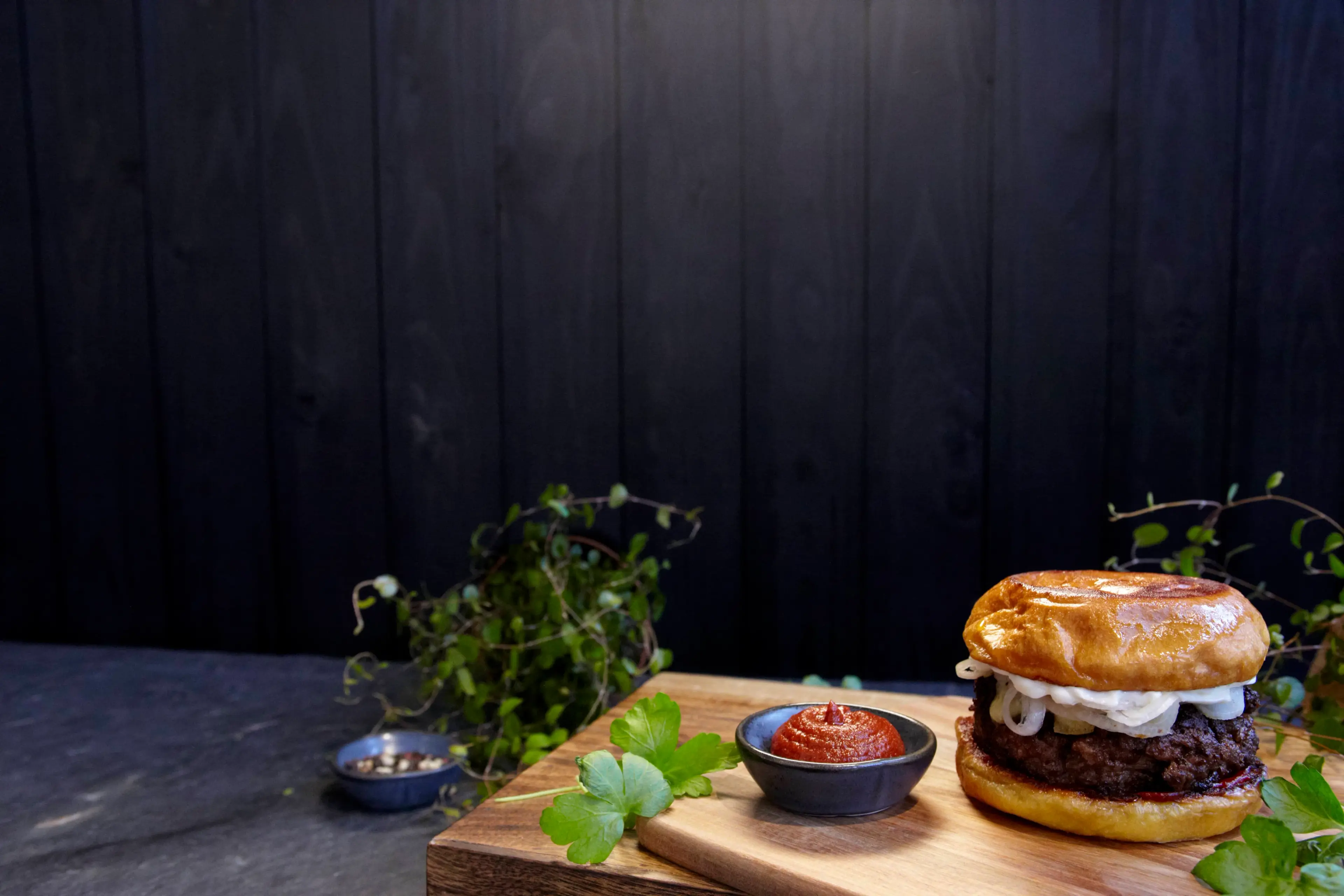 En saftig hamburgare med stekt lök i ett gyllene, mjukt hamburgerbröd står på en skärbräda tillsammans med en skål kryddig rödpaprikasås och färska persiljeblad. Mörk träbakgrund och gröna