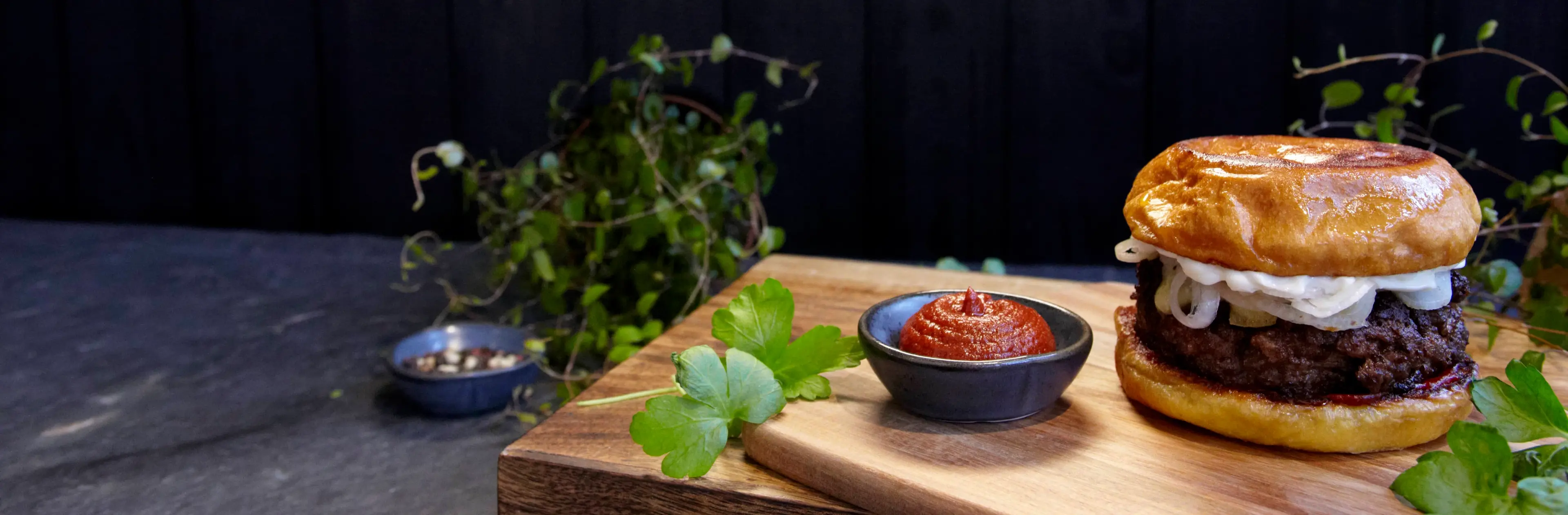 En saftig hamburgare med stekt lök i ett gyllene, mjukt hamburgerbröd står på en skärbräda tillsammans med en skål kryddig rödpaprikasås och färska persiljeblad. Mörk träbakgrund och gröna