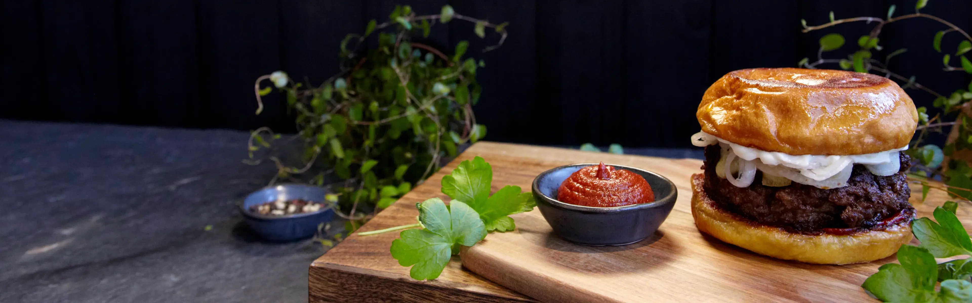En saftig hamburgare med stekt lök i ett gyllene, mjukt hamburgerbröd står på en skärbräda tillsammans med en skål kryddig rödpaprikasås och färska persiljeblad. Mörk träbakgrund och gröna