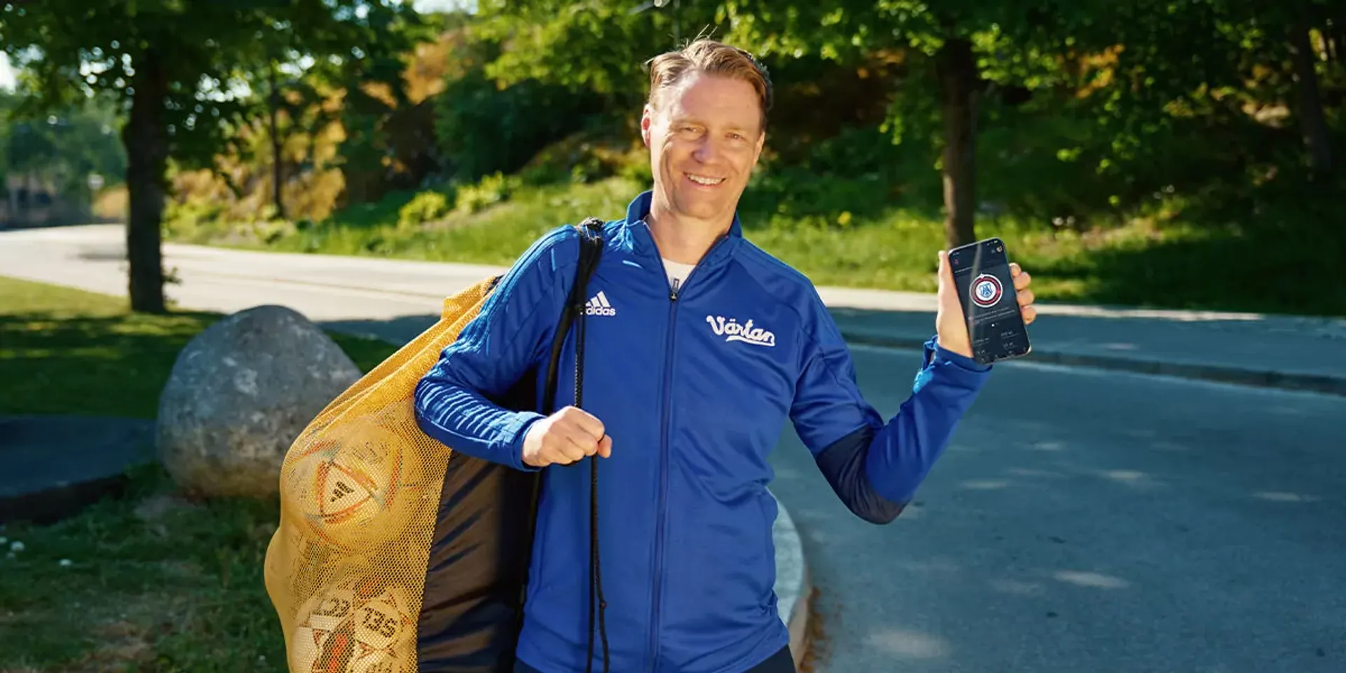 En man i blå träningsjacka bär en nätkasse med fotbollar och visar en mobiltelefon med en app på skärmen. Bakgrunden är en grönskande parkmiljö vid en asfalterad väg.