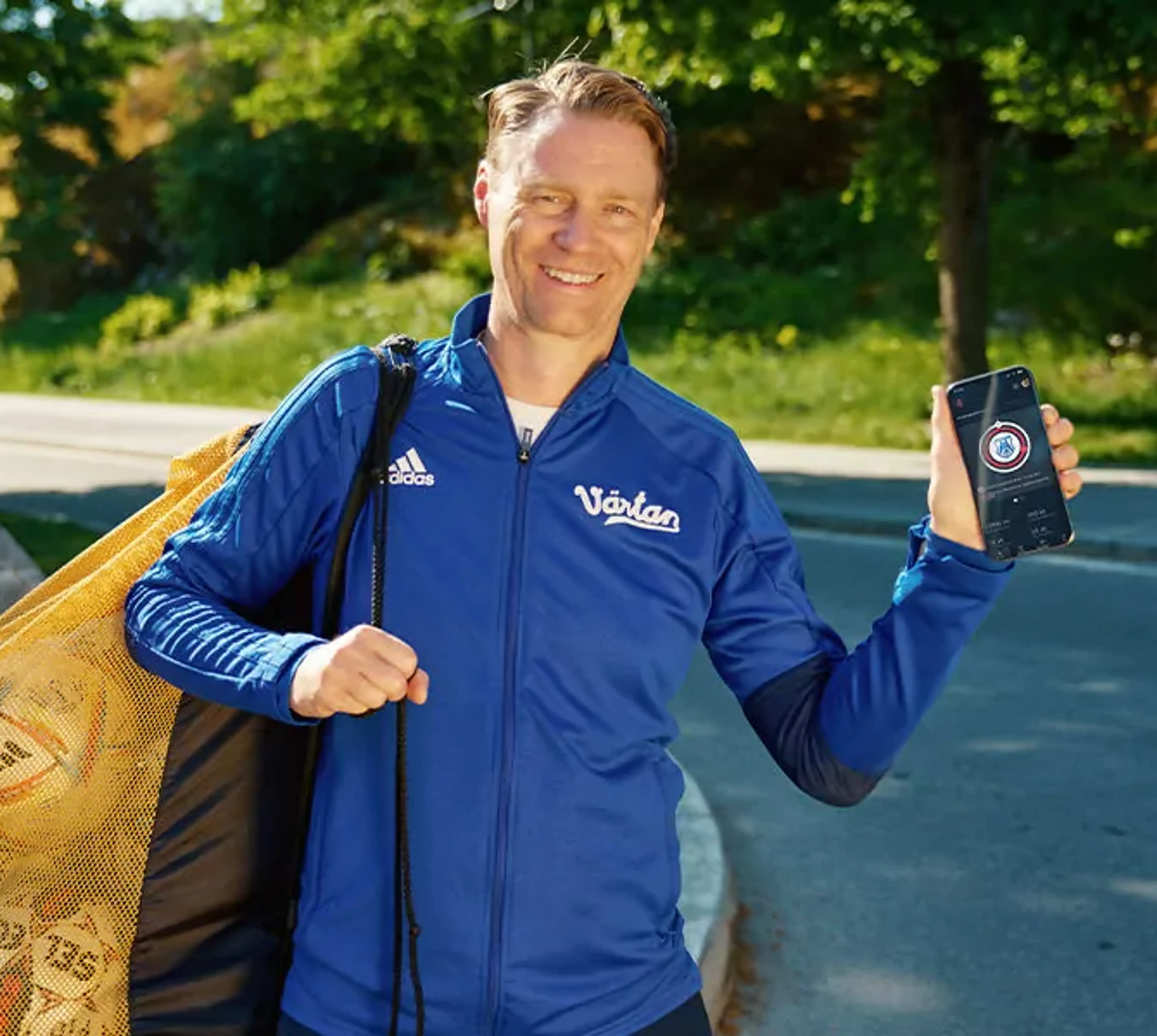 En man i blå träningsjacka bär en nätkasse med fotbollar och visar en mobiltelefon med en app på skärmen. Bakgrunden är en grönskande parkmiljö vid en asfalterad väg.