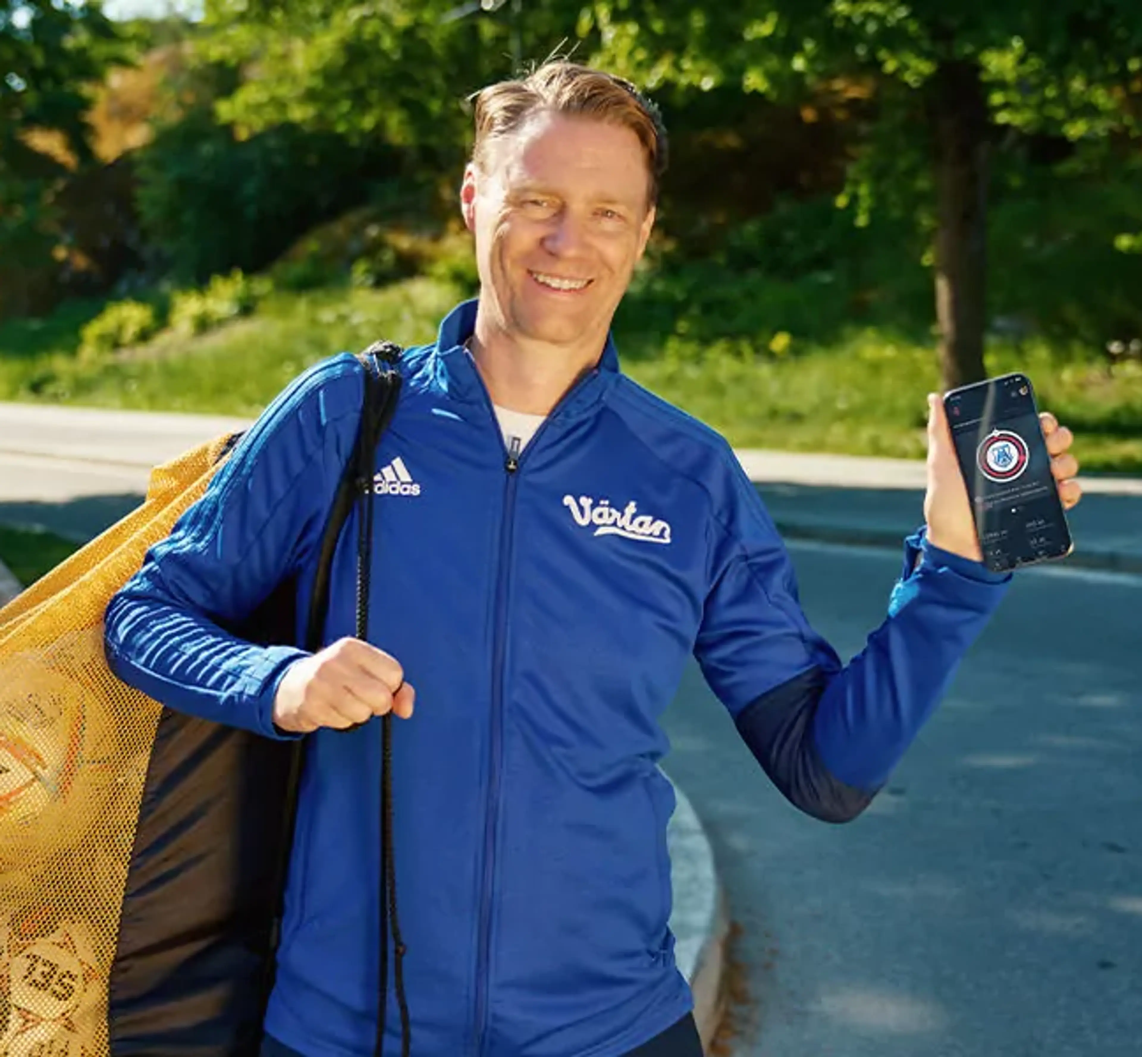 En man i blå träningsjacka bär en nätkasse med fotbollar och visar en mobiltelefon med en app på skärmen. Bakgrunden är en grönskande parkmiljö vid en asfalterad väg.