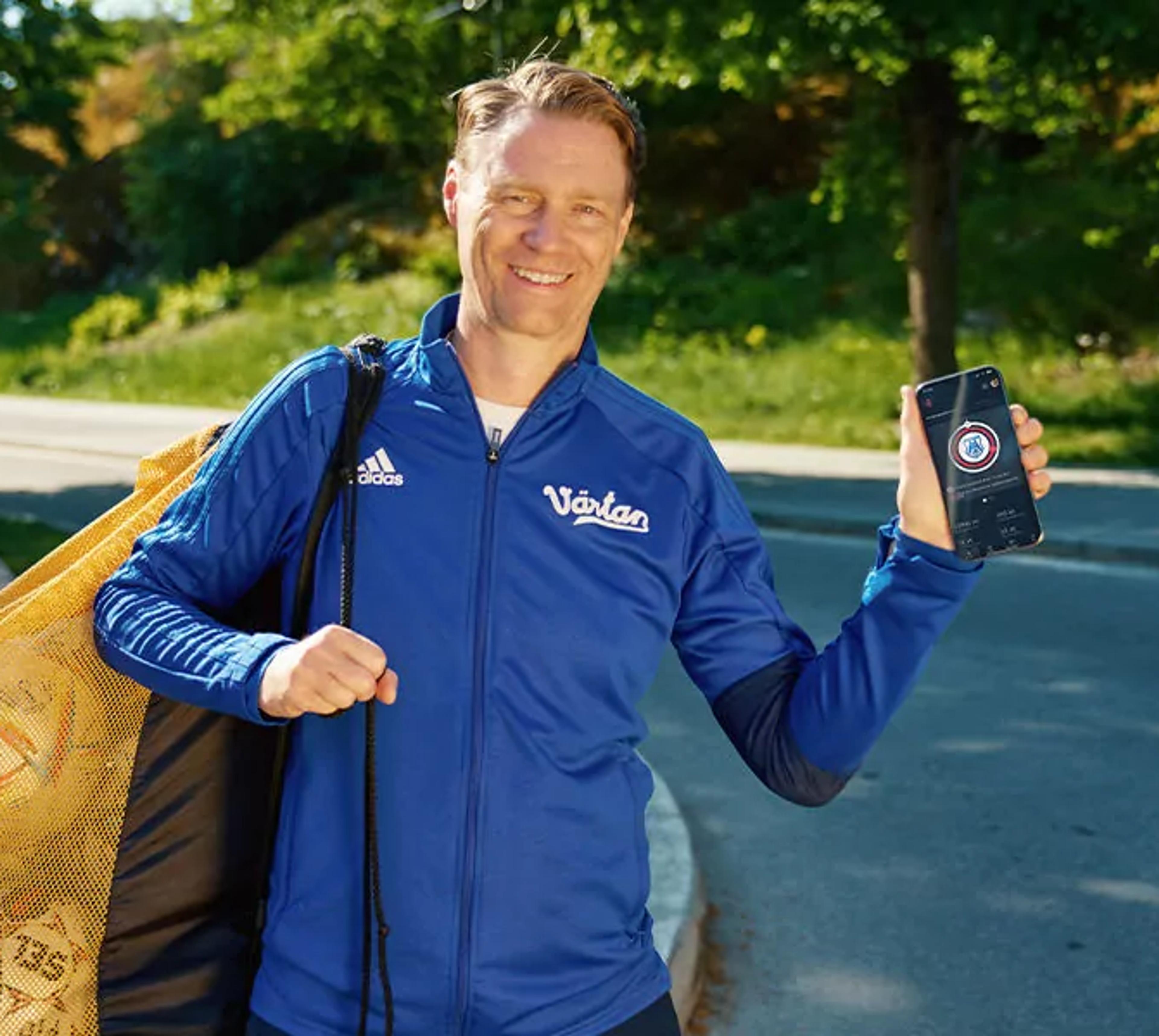En man i blå träningsjacka bär en nätkasse med fotbollar och visar en mobiltelefon med en app på skärmen. Bakgrunden är en grönskande parkmiljö vid en asfalterad väg.