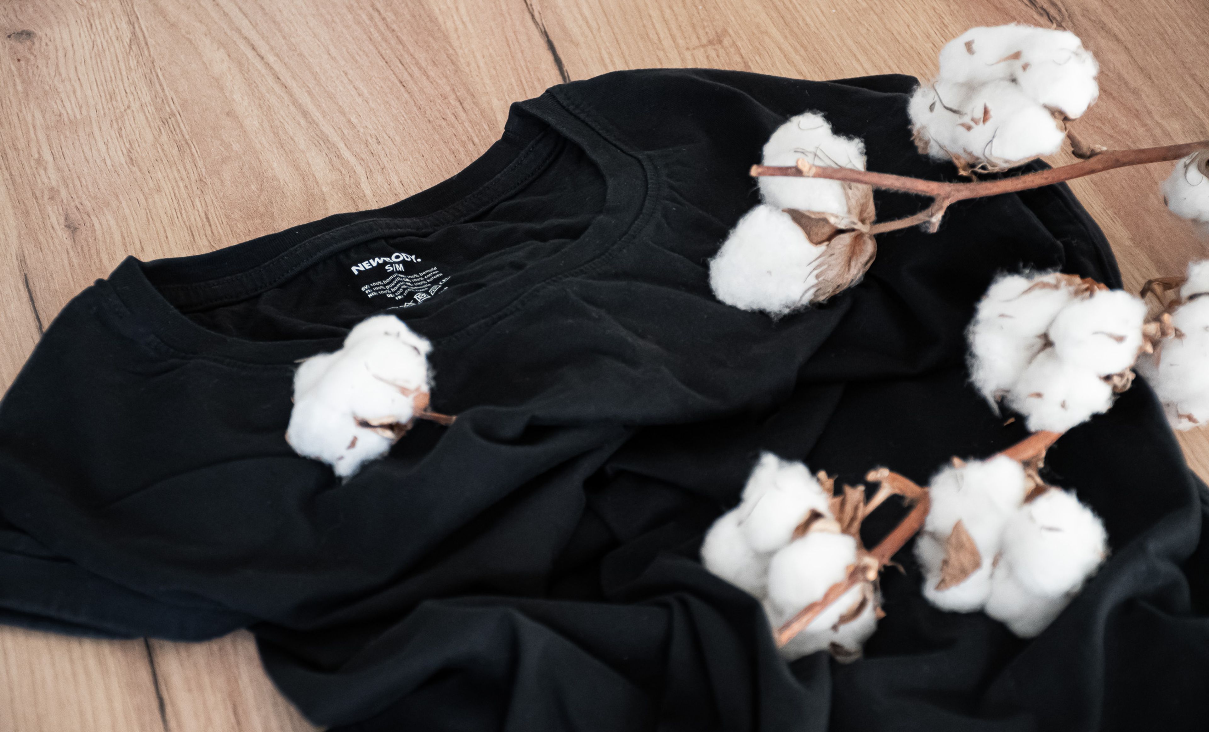 Svart, mjuk t-shirt i bomull ligger tillsammans med några bomullskvistar på ett ljust träunderlag.