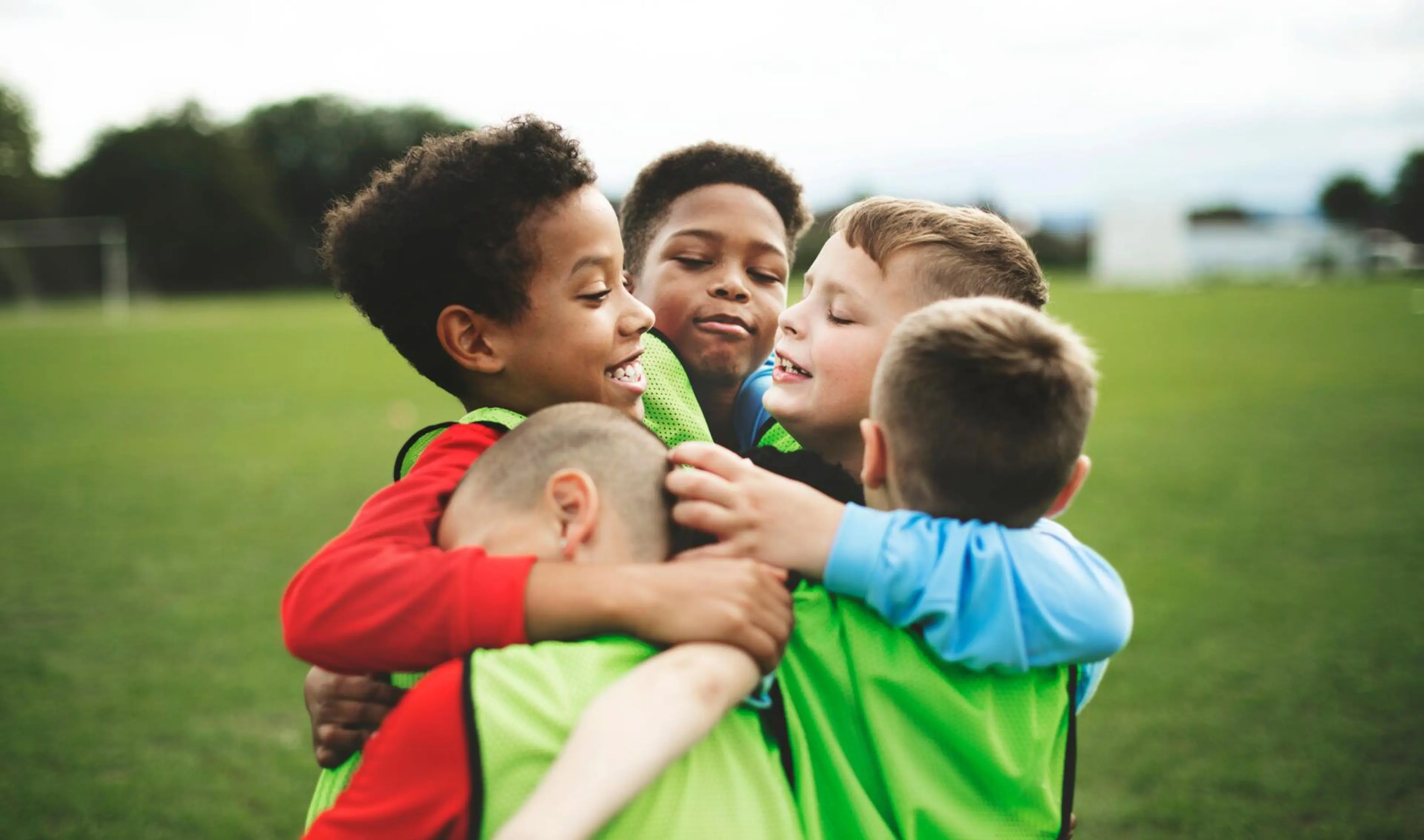 Fem barn i träningsvästar kramas tätt tillsammans på en grön fotbollsplan. Bakgrunden visar en otydlig grönska och ett mål.