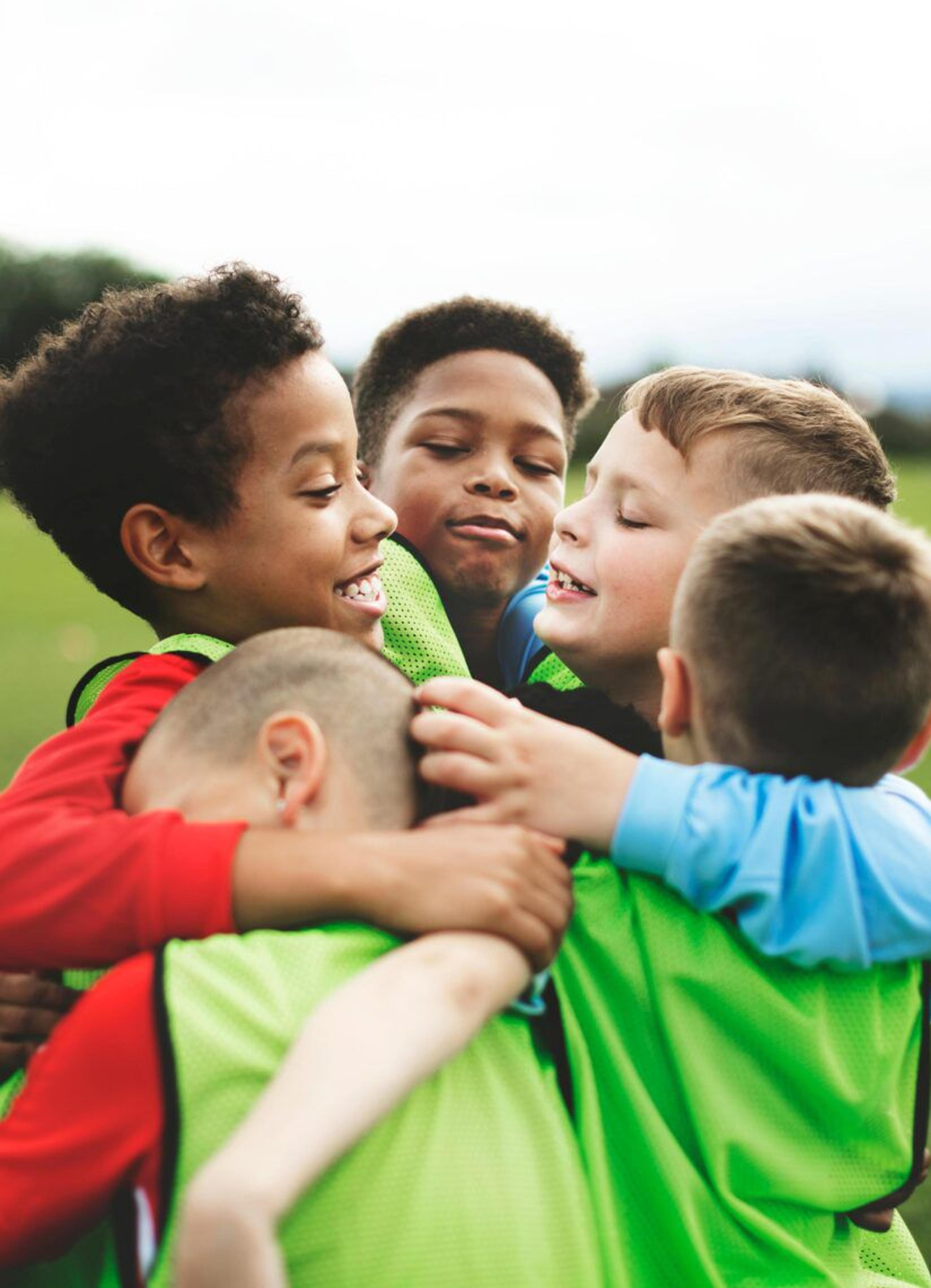 Fem barn i träningsvästar kramas tätt tillsammans på en grön fotbollsplan. Bakgrunden visar en otydlig grönska och ett mål.