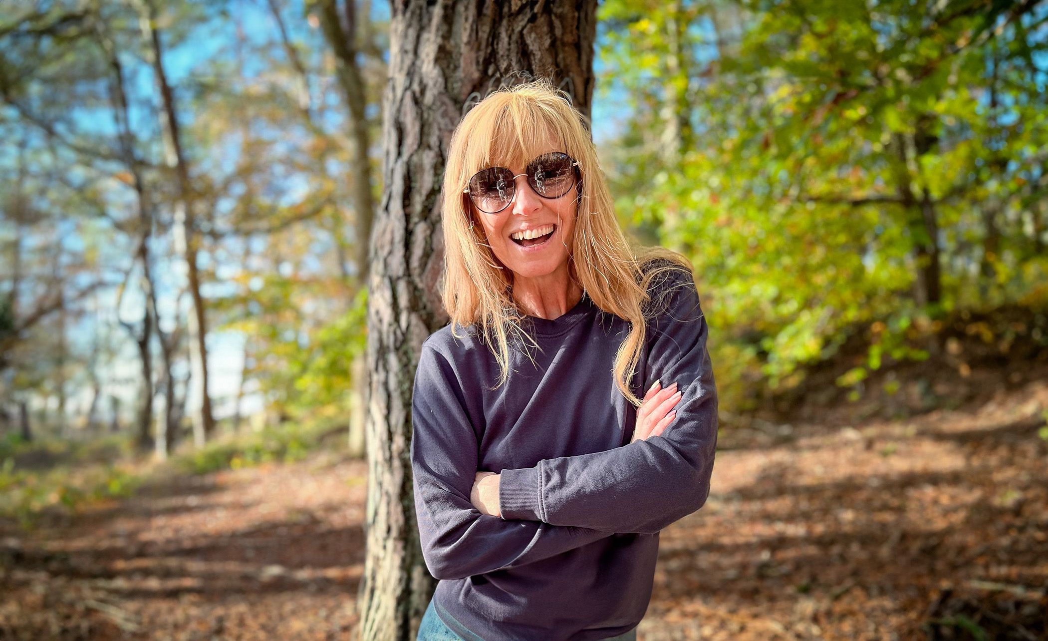 En kvinna med solglasögon och långt blont hår står med armarna i kors framför en trädstam i en skogsmiljö. Hon bär en mörk, långärmad tröja och ler mot kameran.