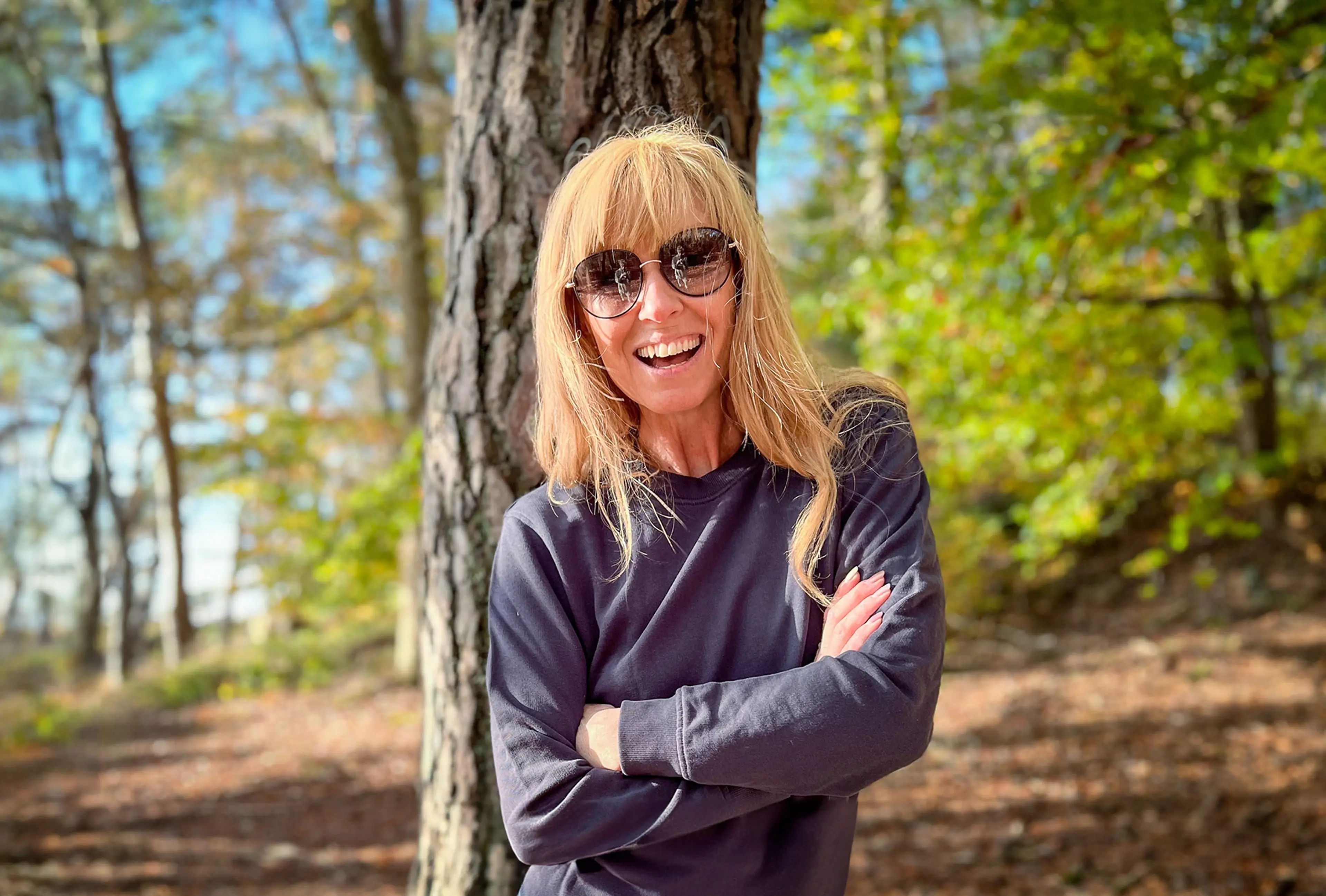 En kvinna med solglasögon och långt blont hår står med armarna i kors framför en trädstam i en skogsmiljö. Hon bär en mörk, långärmad tröja och ler mot kameran.