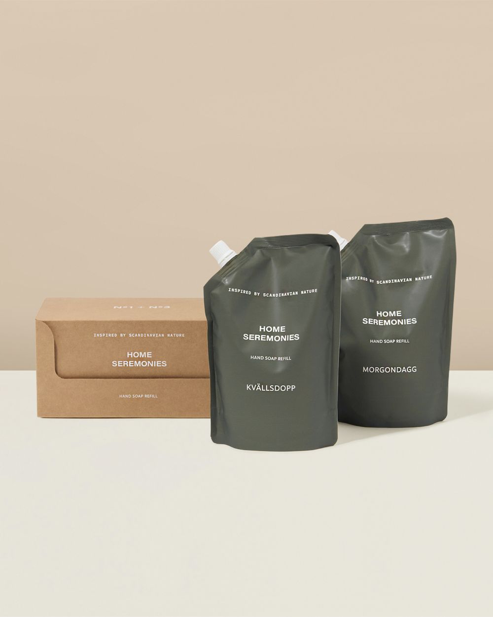 Två påfyllningspåsar för handtvål i mörkgrön färg med texten "Kvällsdopp" och "Morgondagg", tillsammans med en beige kartongförpackning. Enkel och stilren design inspirerad av skandinavisk natur.