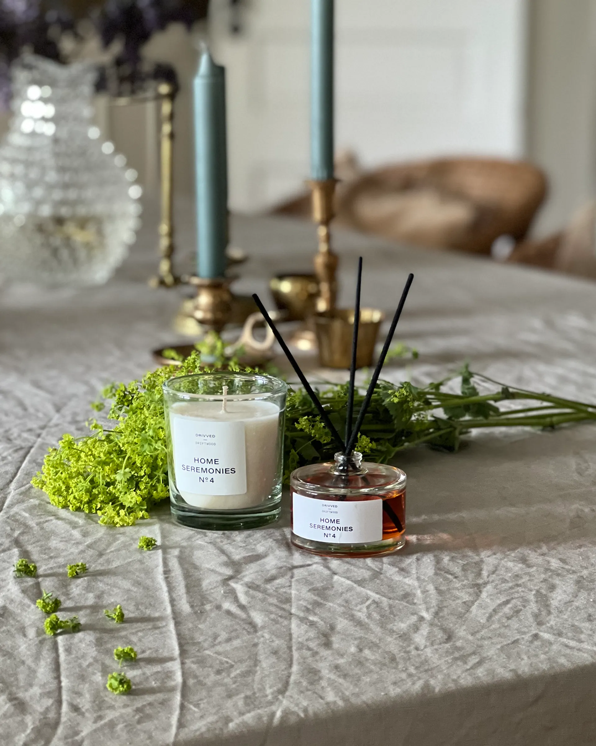 Ett doftljus i glas och en glasflaska med doftpinnar, båda med etikett och doften drivved, placerade på ett bord med gröna växtdetaljer och blå ljus i bakgrunden.