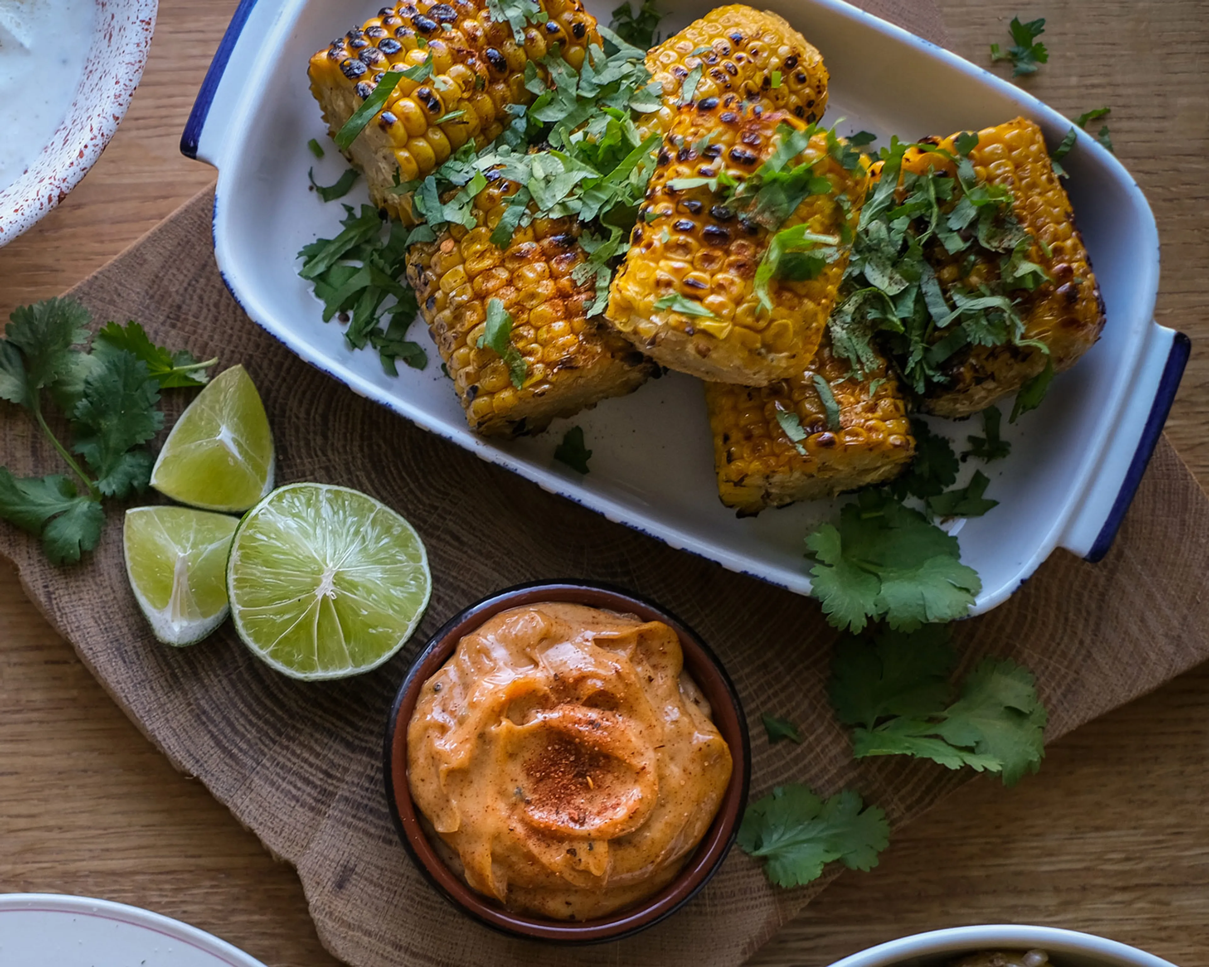 Grillade majskolvar med chilipulver och koriander i en ugnsform, serverade med en krämig, kryddig dipp och limeklyftor på ett träfat.