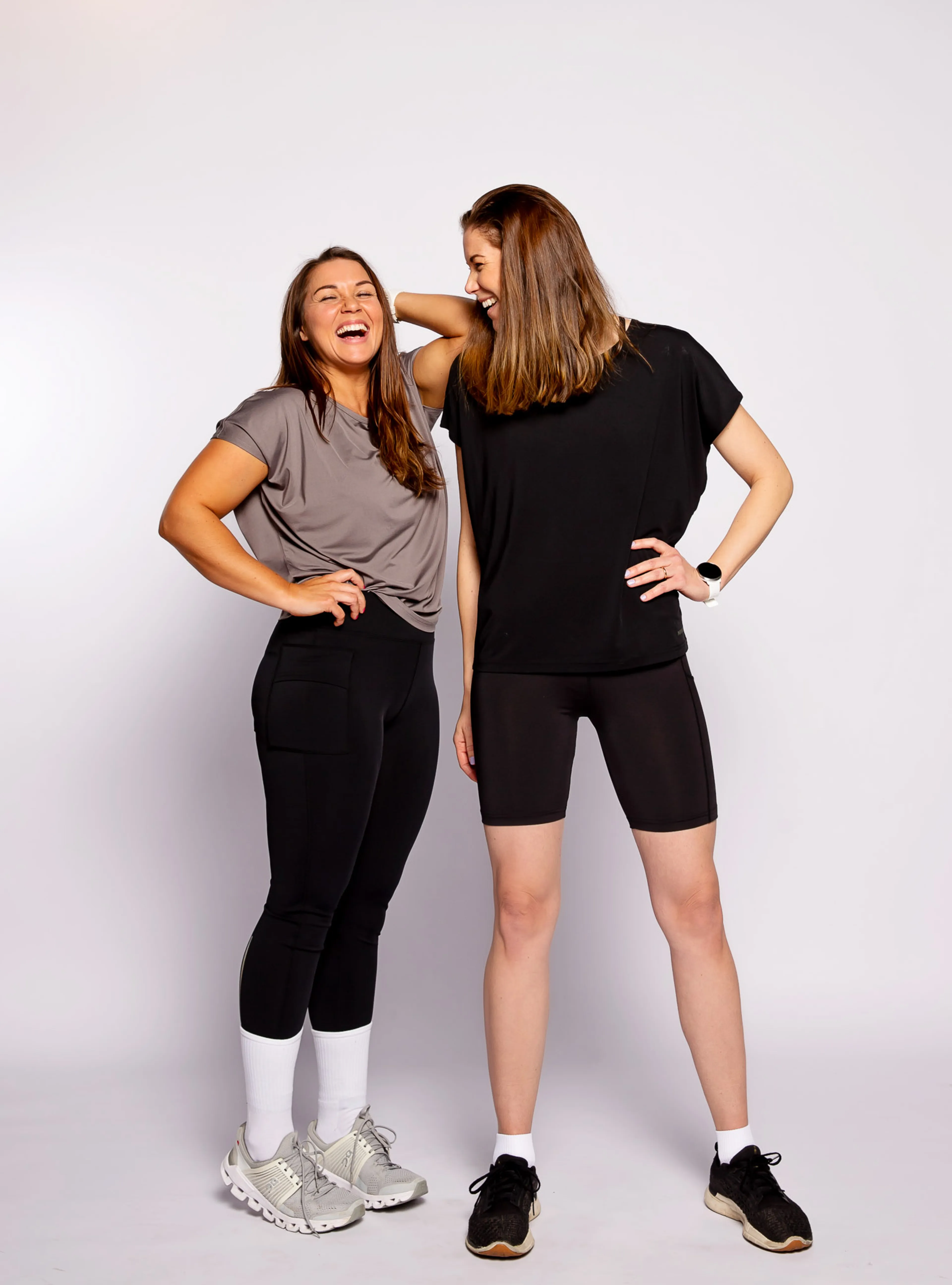 Två kvinnor i sportiga kläder, där den ena bär en grå t-shirt och svarta tights med fickor och den andra en svart t-shirt och svarta cykelbyxor. Båda har sneakers och ser glada ut.