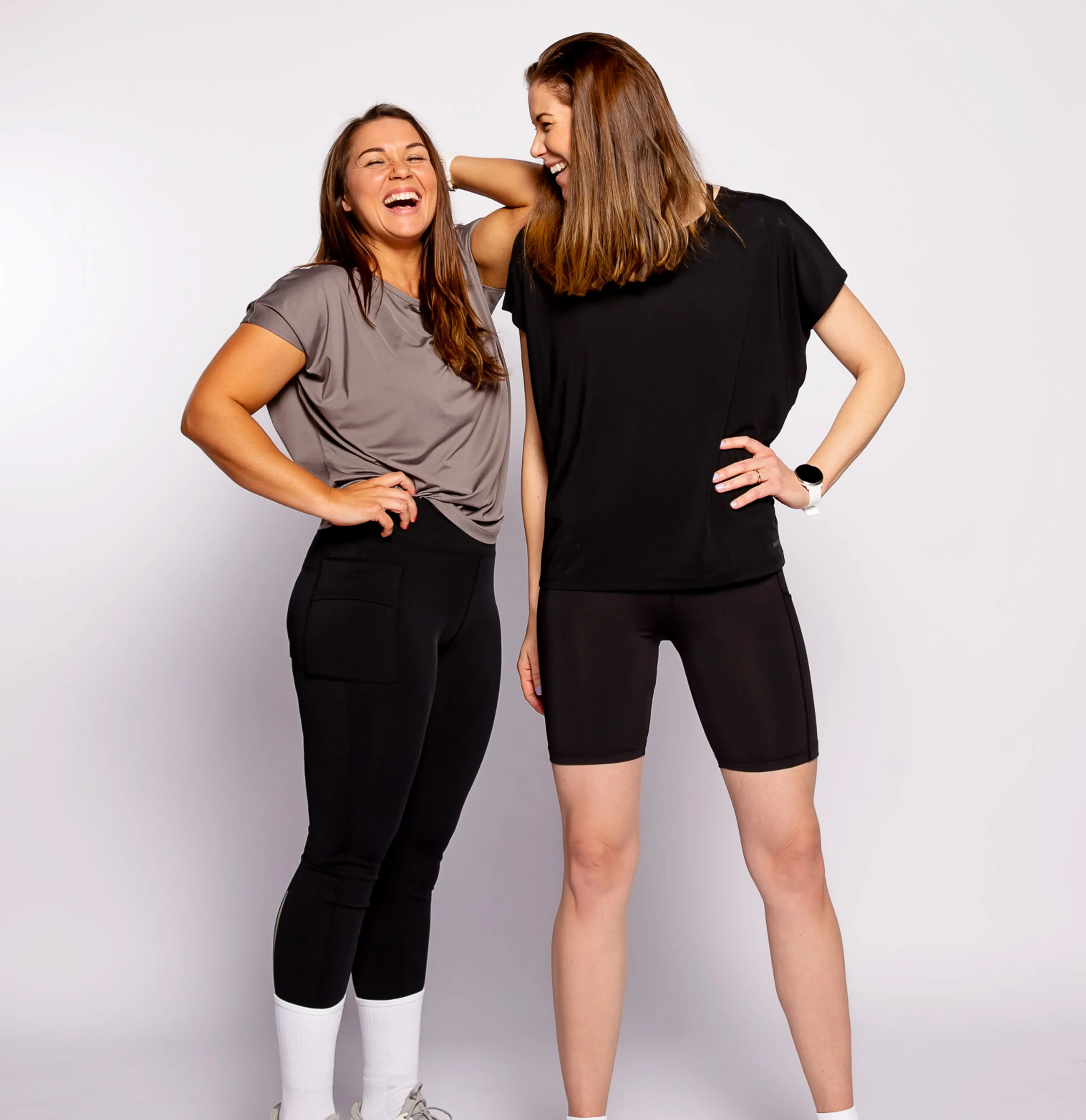 Två kvinnor i sportiga kläder, där den ena bär en grå t-shirt och svarta tights med fickor och den andra en svart t-shirt och svarta cykelbyxor. Båda har sneakers och ser glada ut.