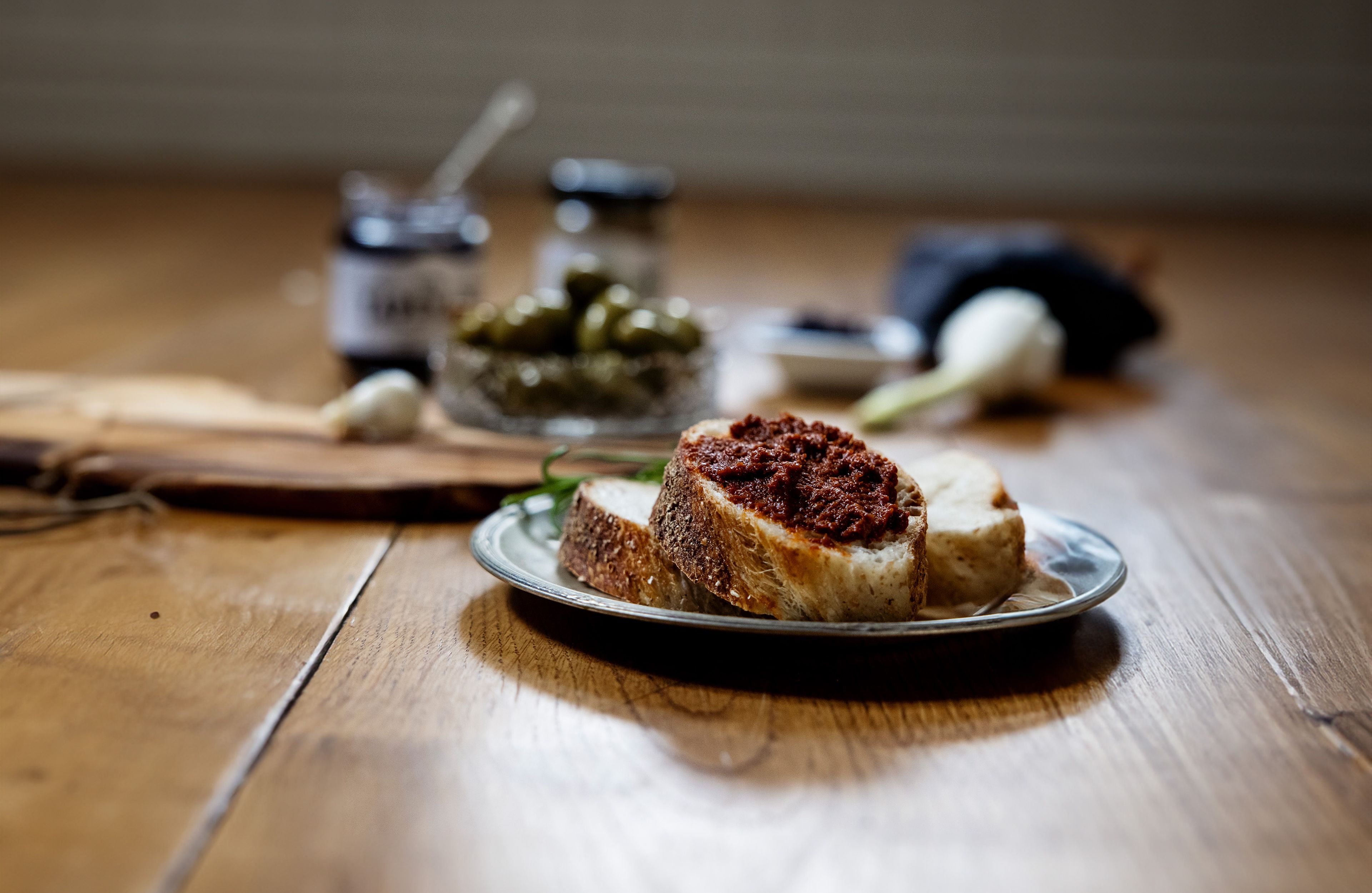 Skivor av bröd med en mörkröd tapenade bredvid en skål med gröna oliver och andra ingredienser på ett träbord.