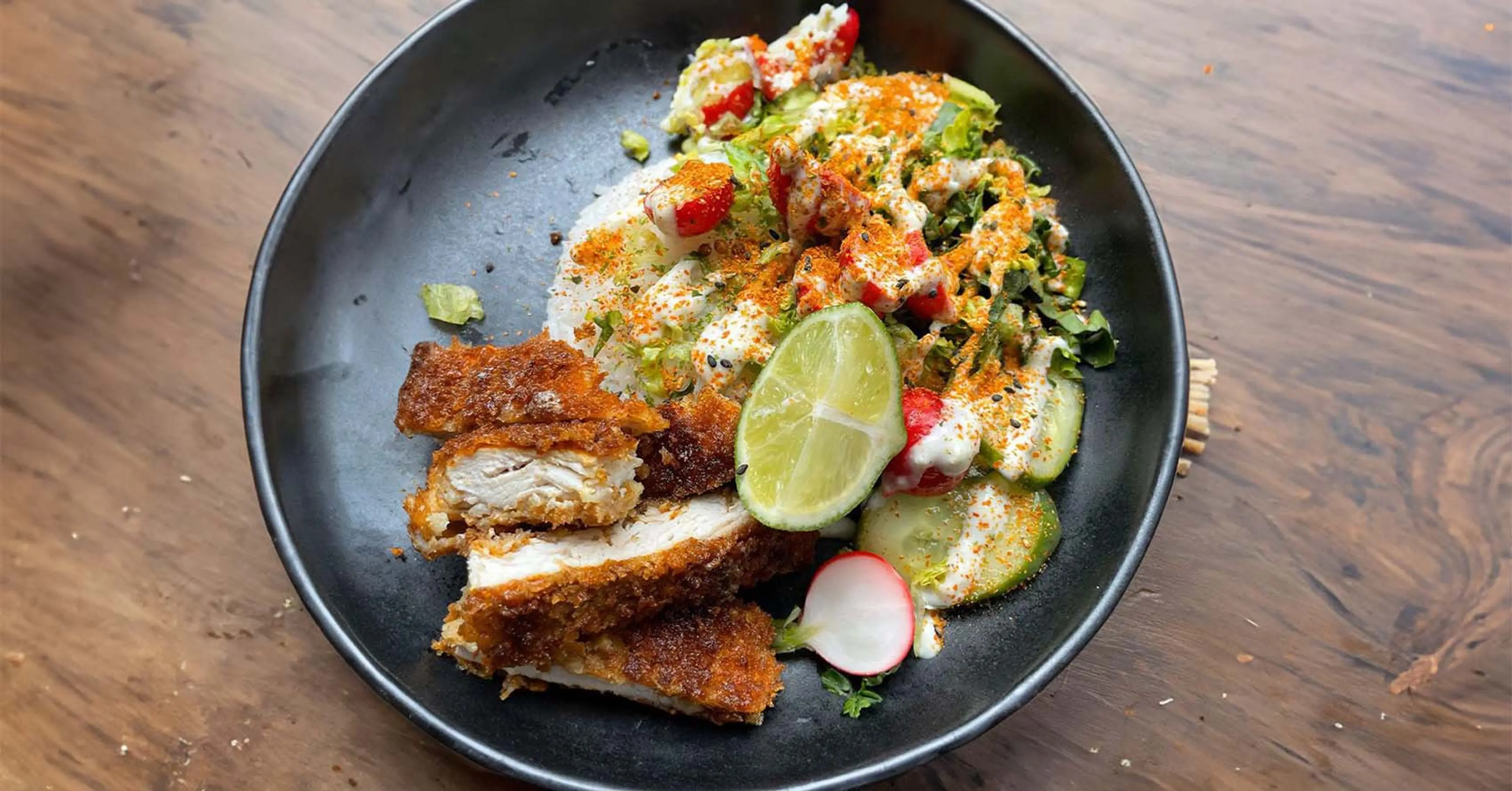 En tallrik med krispig, panerad kycklingschnitzel serverad med färsk sallad, skivade rädisor, gurka och en limeklyfta, toppad med en krämig sås och kryddor.