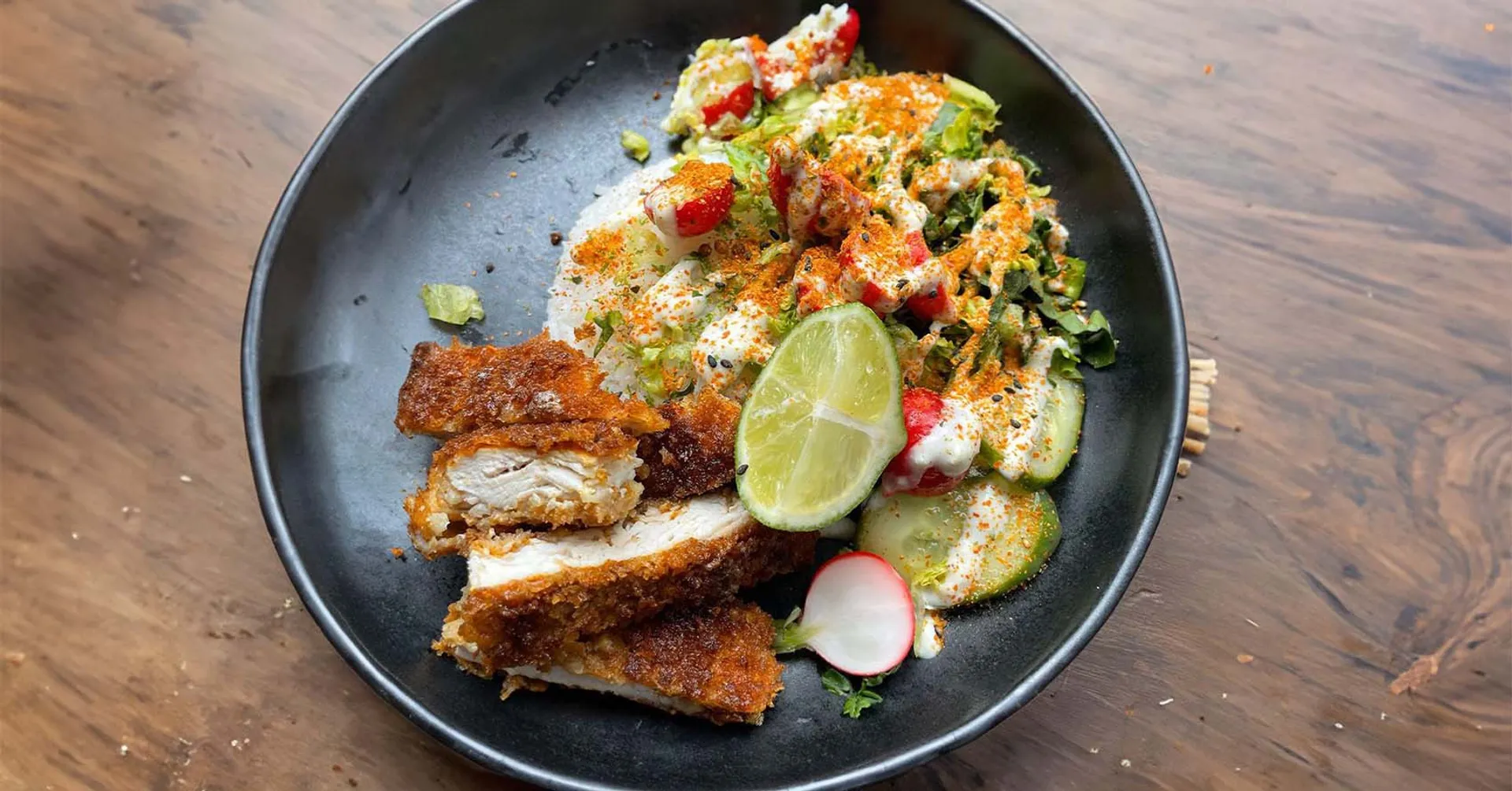 En tallrik med krispig, panerad kycklingschnitzel serverad med färsk sallad, skivade rädisor, gurka och en limeklyfta, toppad med en krämig sås och kryddor.