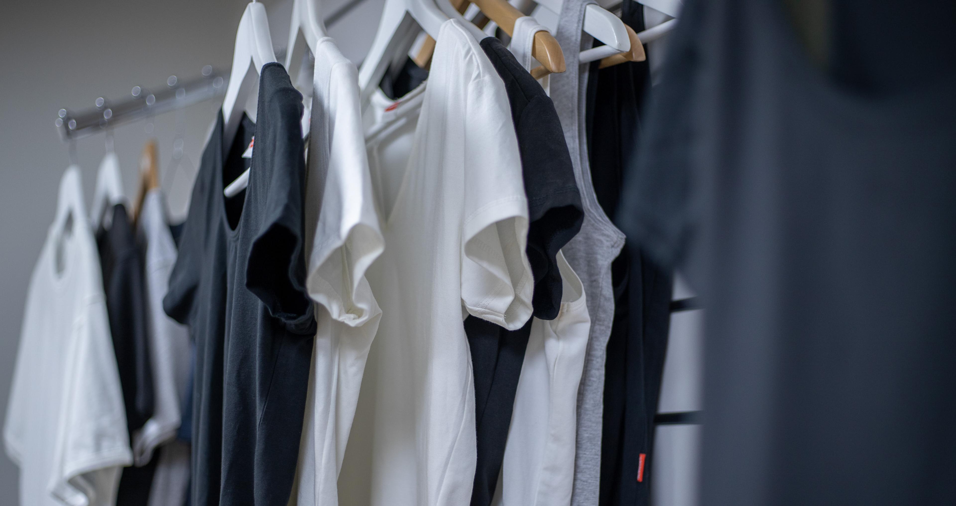 Svarta, vita och grå t-shirts och linnen hänger prydligt på galgar i rad. Kläderna har en enkel och klassisk design i mjukt material.