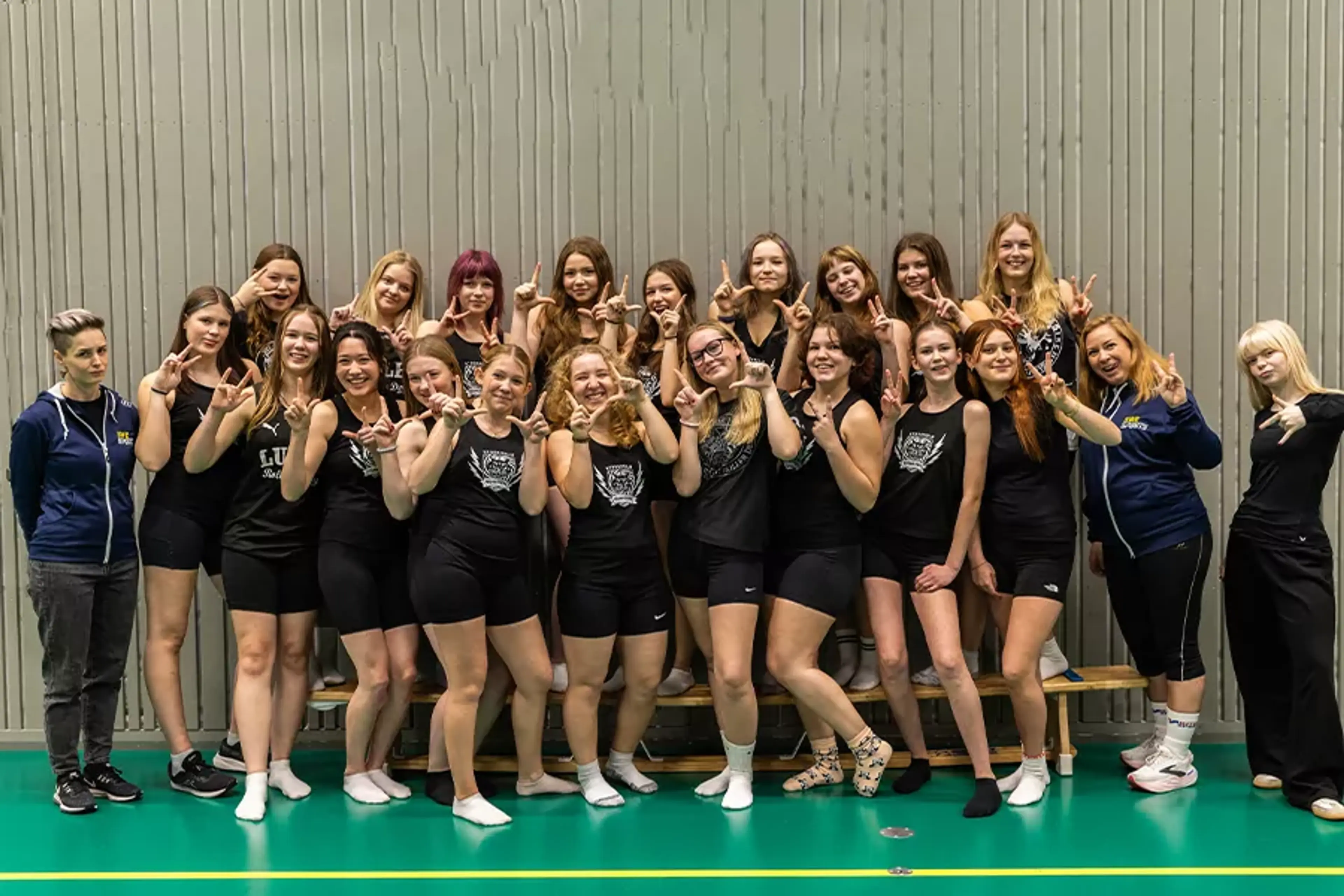 En grupp unga tjejer i sportkläder står samlade i en idrottshall och visar tecken med händerna. Två ledare i sportkläder finns också med på bilden.