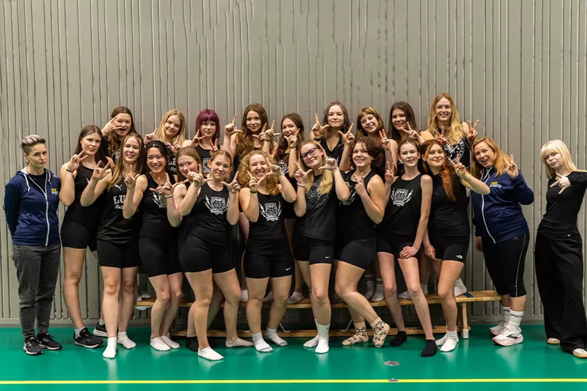 En grupp unga tjejer i sportkläder står samlade i en idrottshall och visar tecken med händerna. Två ledare i sportkläder finns också med på bilden.