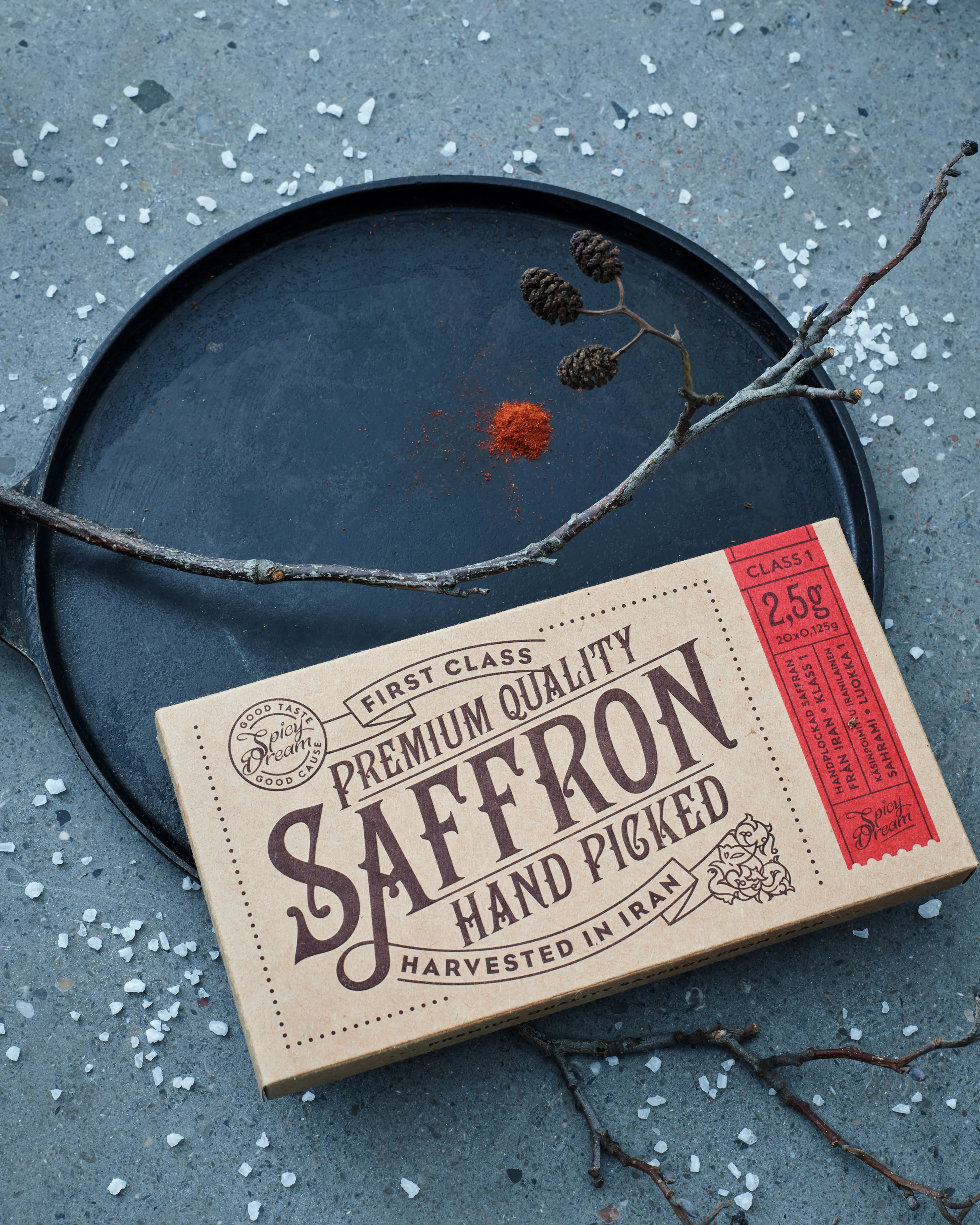 Ett paket med handplockad saffran av premiumkvalitet i enkelt pappersomslag. En svart rund bricka med en liten hög saffran och några tunna grenar ligger intill.