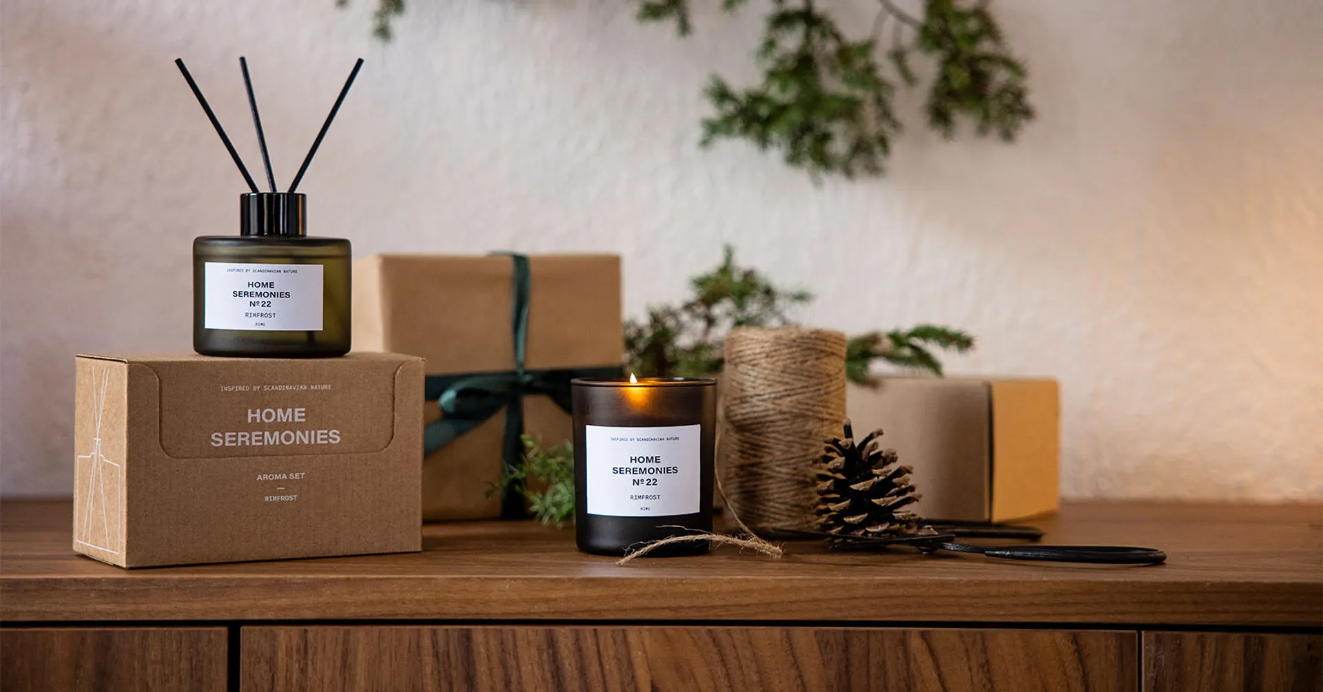 Ett doftset med rökelsepinnar i en grågrön glasflaska och ett tänd doftljus i matchande mörkt glas, båda märkta med "Rimfrost". Paket och julpynt som kottar, snöre och