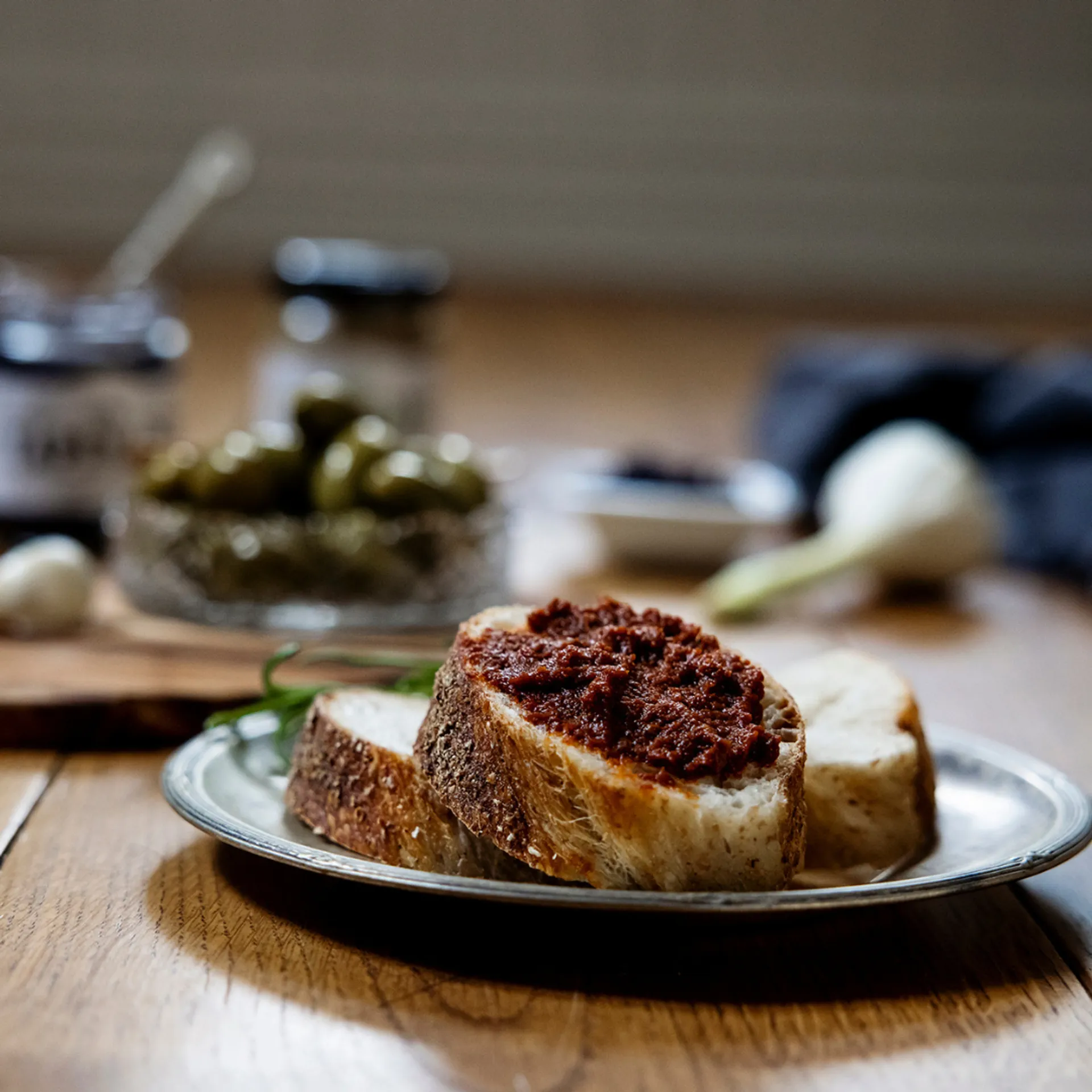 Tre skivor bröd på ett fat, varav en är bred med en krämig röra med djup röd färg. I bakgrunden syns burkar, en skål med gröna oliver och en vitlök.