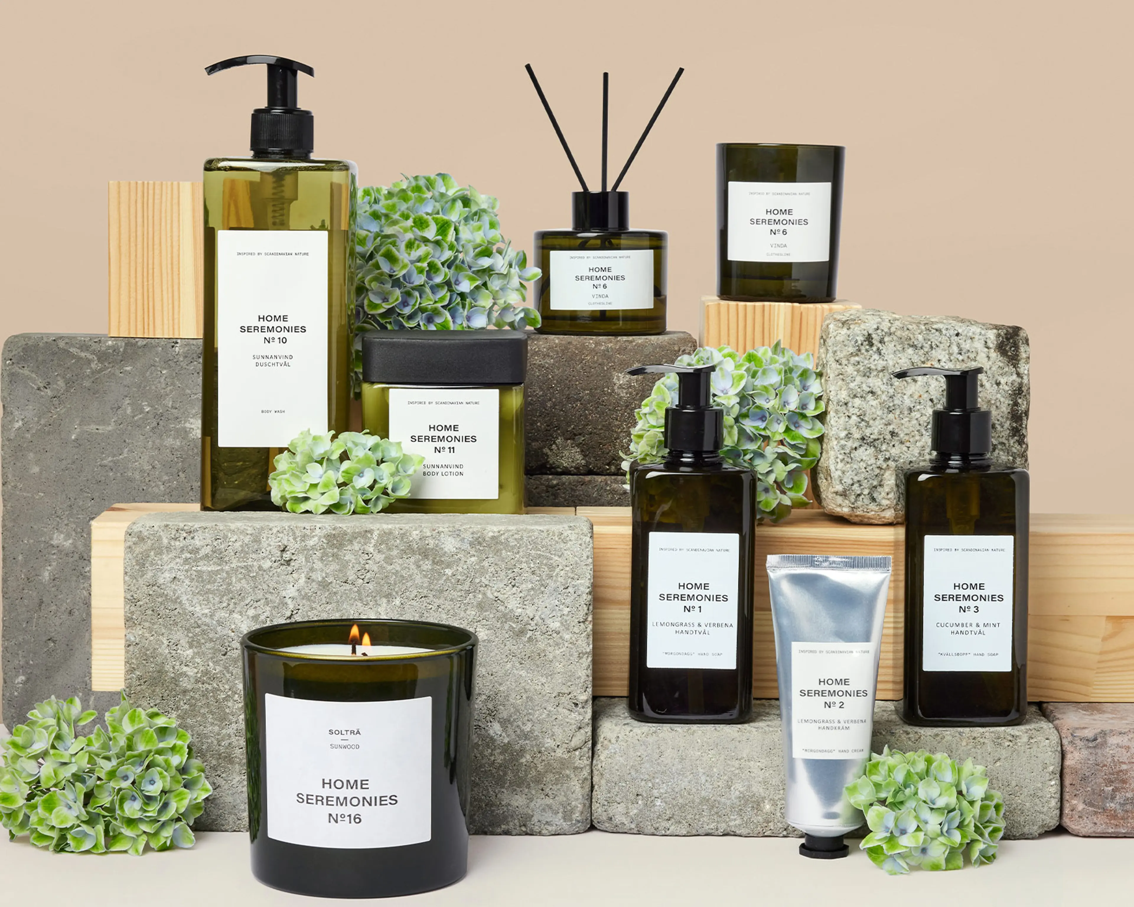 Ett urval av doftljus, handtvålar, handkräm och kroppslotion i gröna glasflaskor och tuber, arrangerade med gråa stenblock och gröna blommor. Produkterna har enkla, vita etiketter med svart text